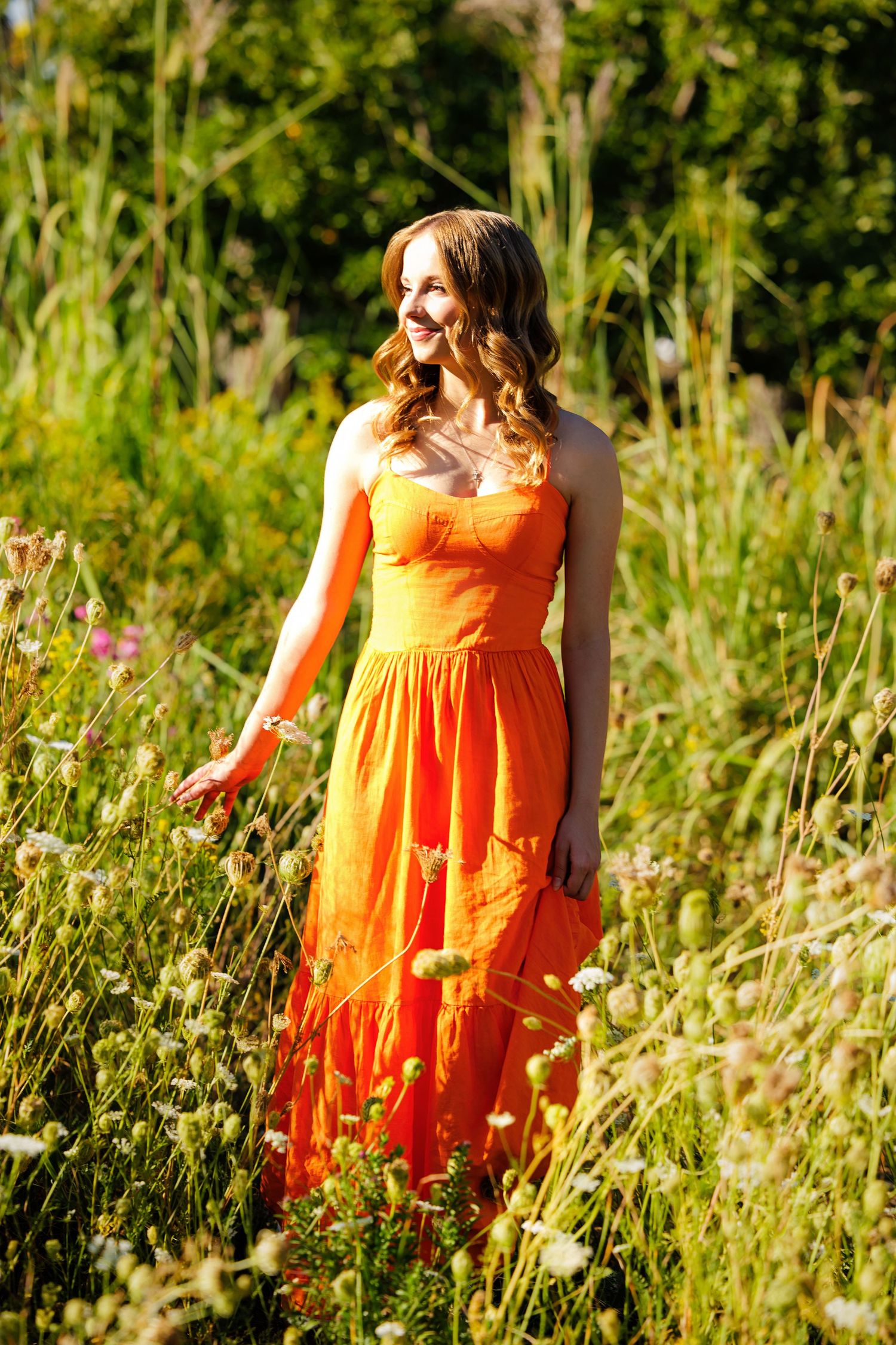Young woman in vibrant orange sundress standing in a wildflower meadow, smiling in golden sunlight in Littleton Colorado