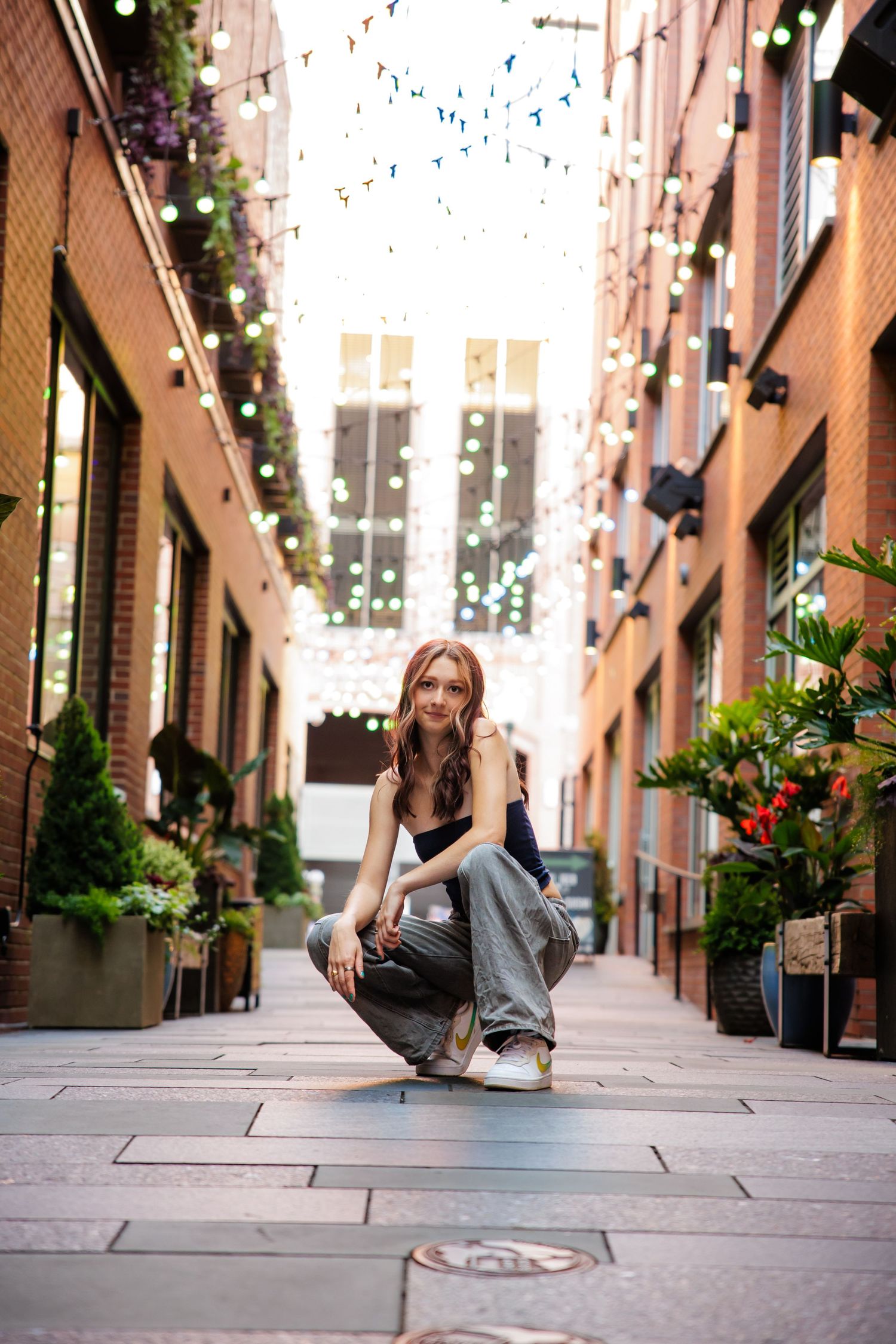 Young woman crouching in decorative alley with string lights and brick walls for Parker CO Urban Senior photos