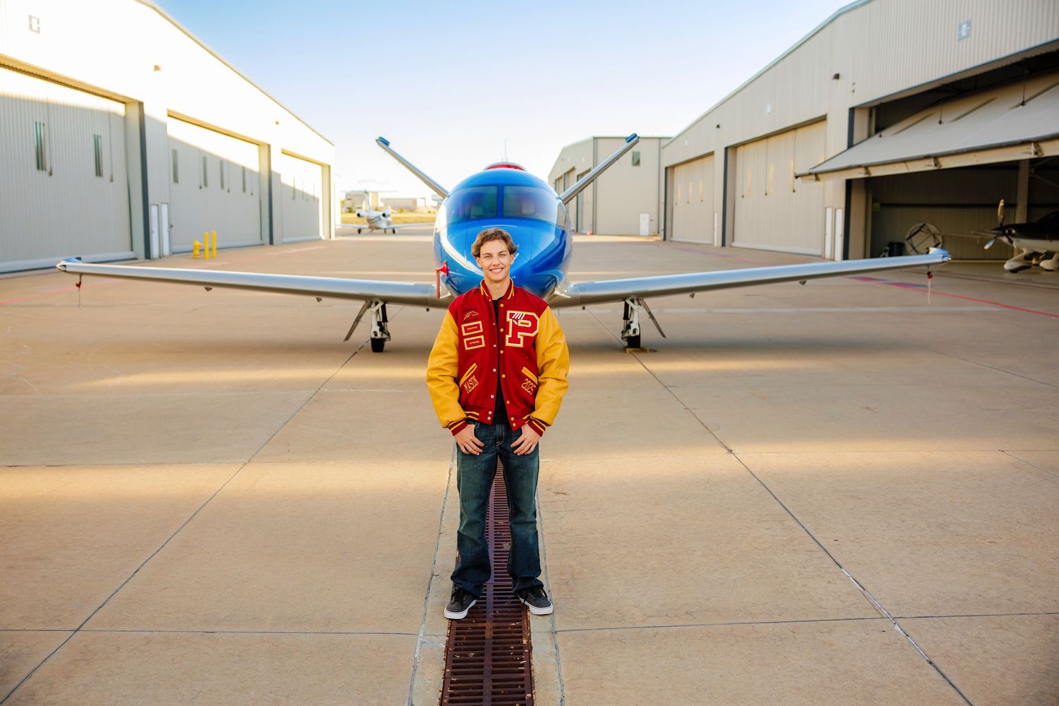 Teen in red varsity jacket posing with arms spread in front of a blue aircraft outdoors.
