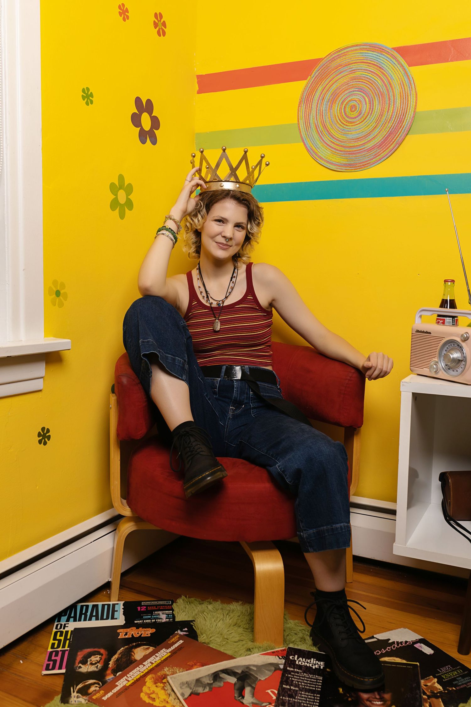 Person wearing gold crown sits in red chair in retro yellow room with vintage magazines posing for senior photos downtown Denver studio