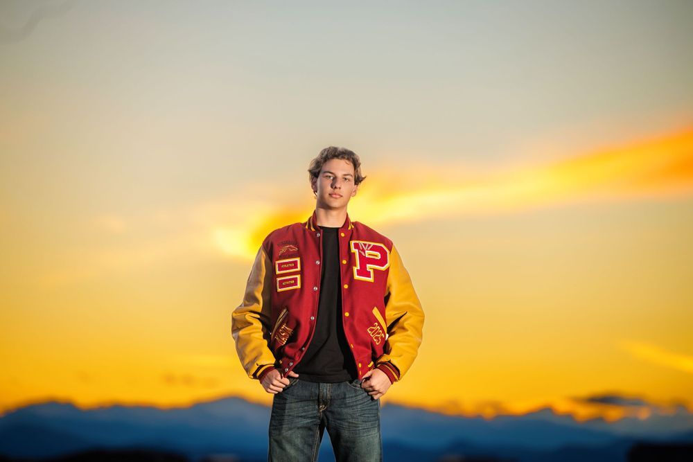 Young man in red and gold letterman jacket standing against a vibrant sunset sky with mountain silhouettes. Sunset Colorado Senior Portrait