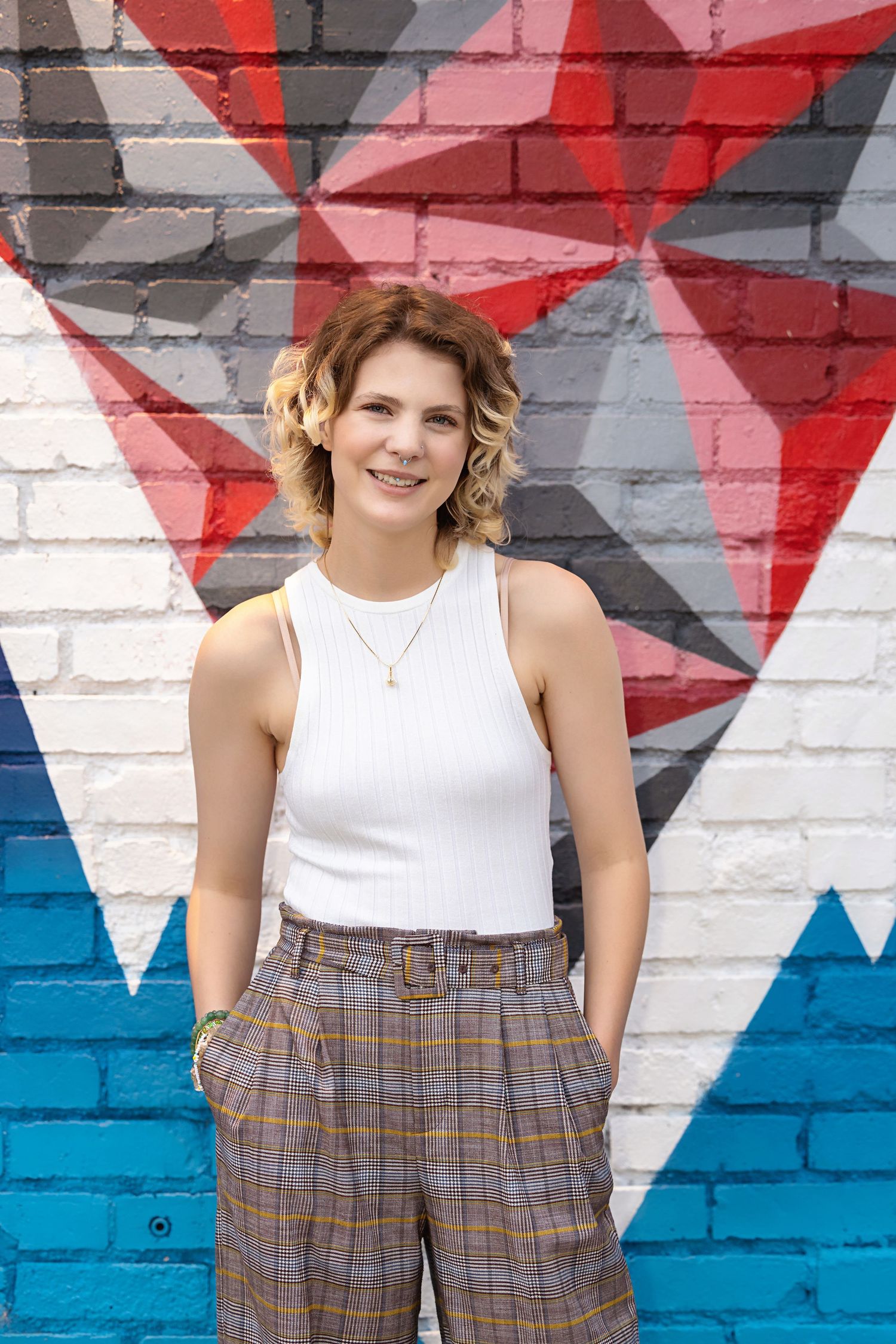 Smiling young person in white tank top and belted plaid pants before colorful geometric brick mural. Denver Colorado Senior Portrait