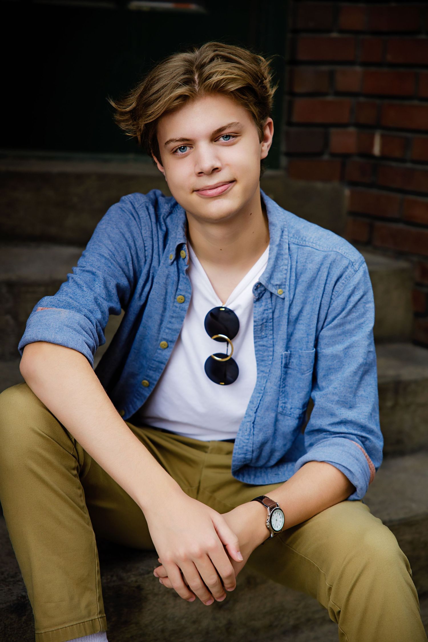 Smiling teen boy in blue shirt and khakis sits on brick steps wearing sunglasses. Parker Colorado Urban Senior Portrait