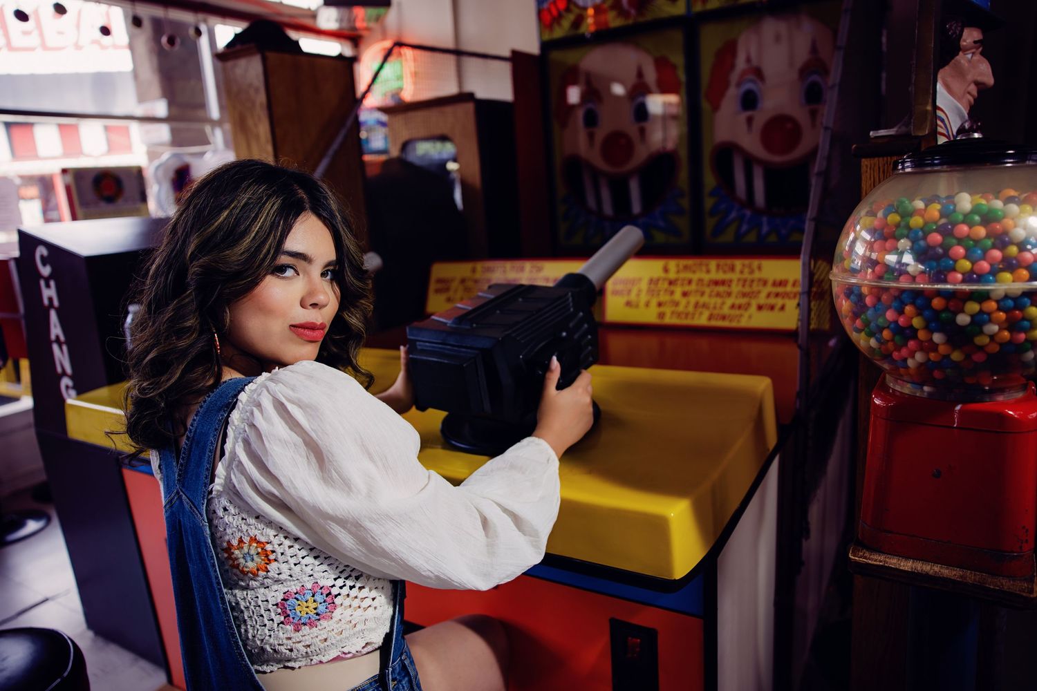 Young woman in denim overalls and white blouse plays arcade shooting game at colorful amusement venue for Aurora Colorado Senior pictures