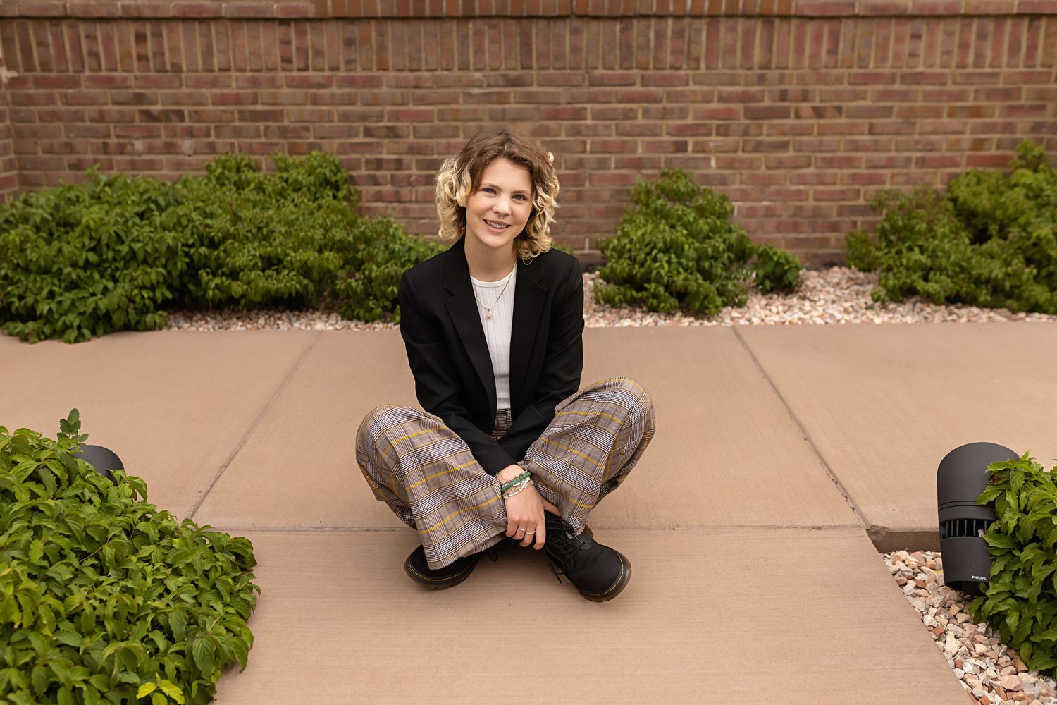 Smiling Parker Colorado LGBTQ friendly images of teen in black blazer and plaid pants sits cross-legged on pavement beside green shrubs.