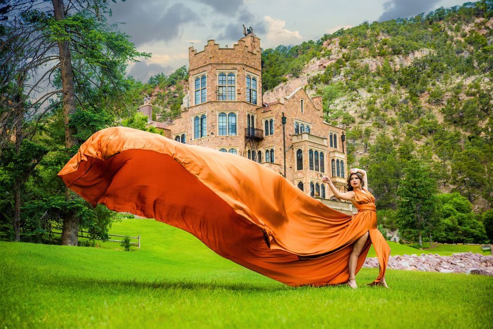 Teen in vibrant orange flying dress with fabric soaring in breeze before historic stone castle Colorado Senior photo session