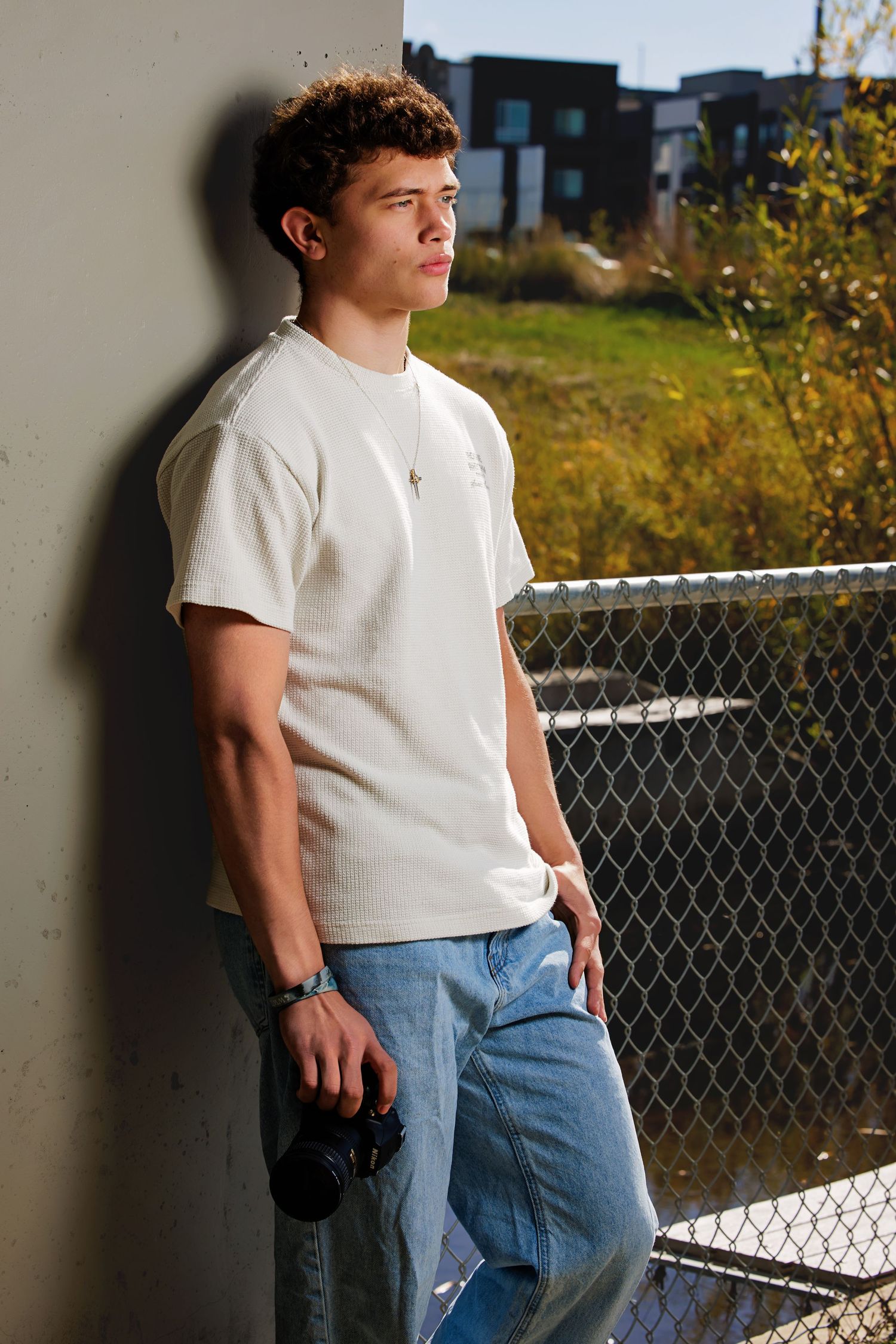Young man in white t-shirt and jeans leaning against wall holding camera outdoors near chain-link fence.