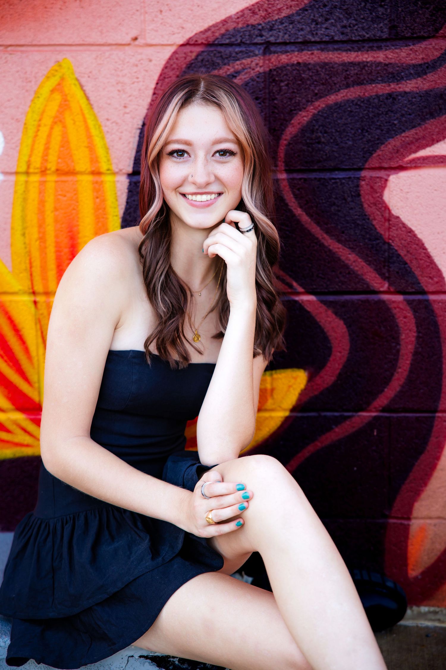 Cheerful young woman in black dress sitting against vibrant painted graffiti wall backdrop for Parker Colorado Senior pictures