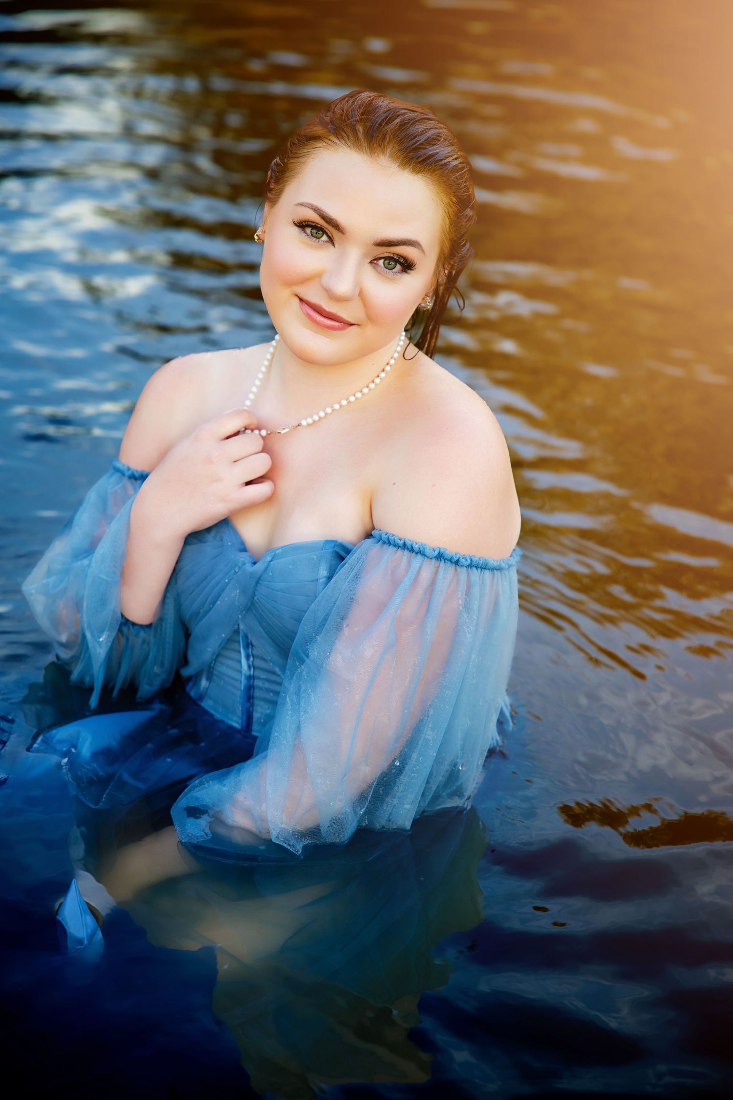 Woman in blue off-shoulder gown wearing pearl necklace sitting in water at sunset. Modern senior photos Parker Colorado