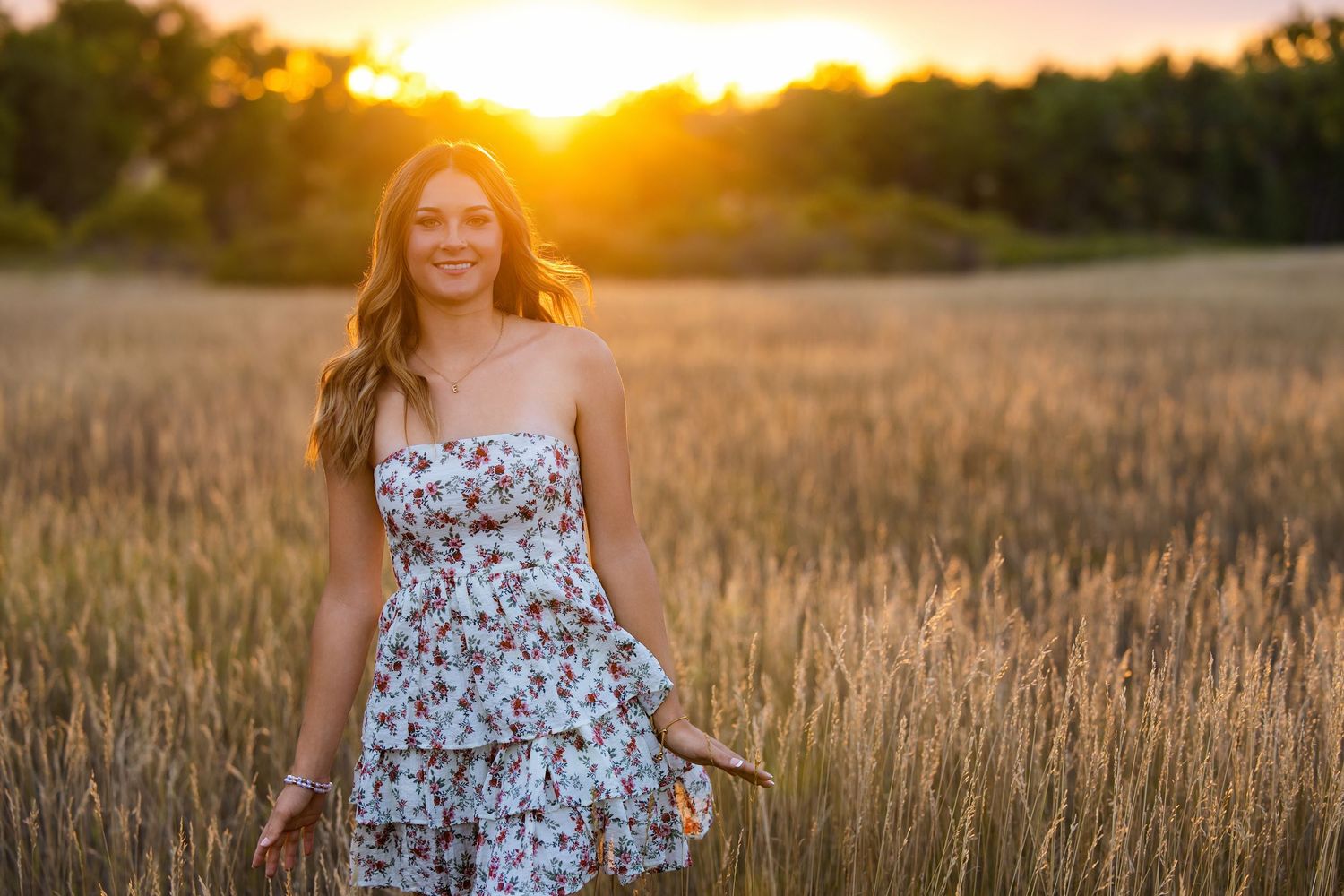 Young woman in floral sundress standing in golden wheat field at sunset.