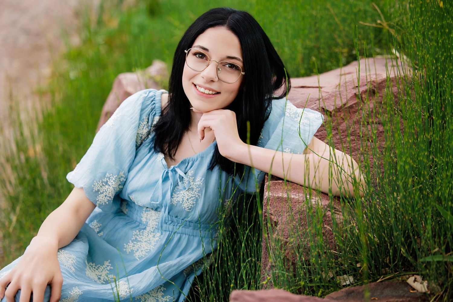 Young woman with dark hair and glasses in a blue floral dress sitting outdoors on rocks surrounded by green grass.