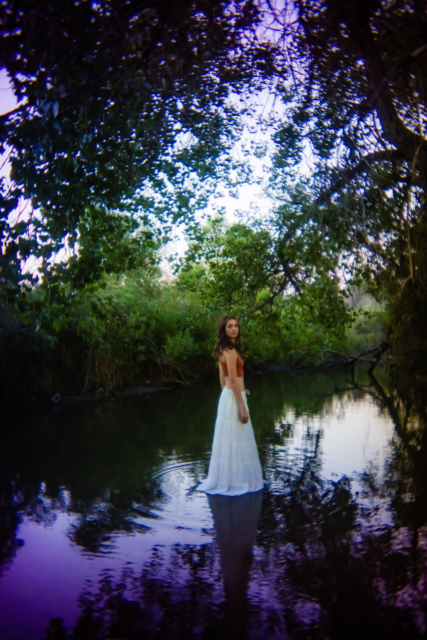 Woman in white backless gown standing in a serene forest pond surrounded by lush green trees for nature Parker Colorado Senior pictures