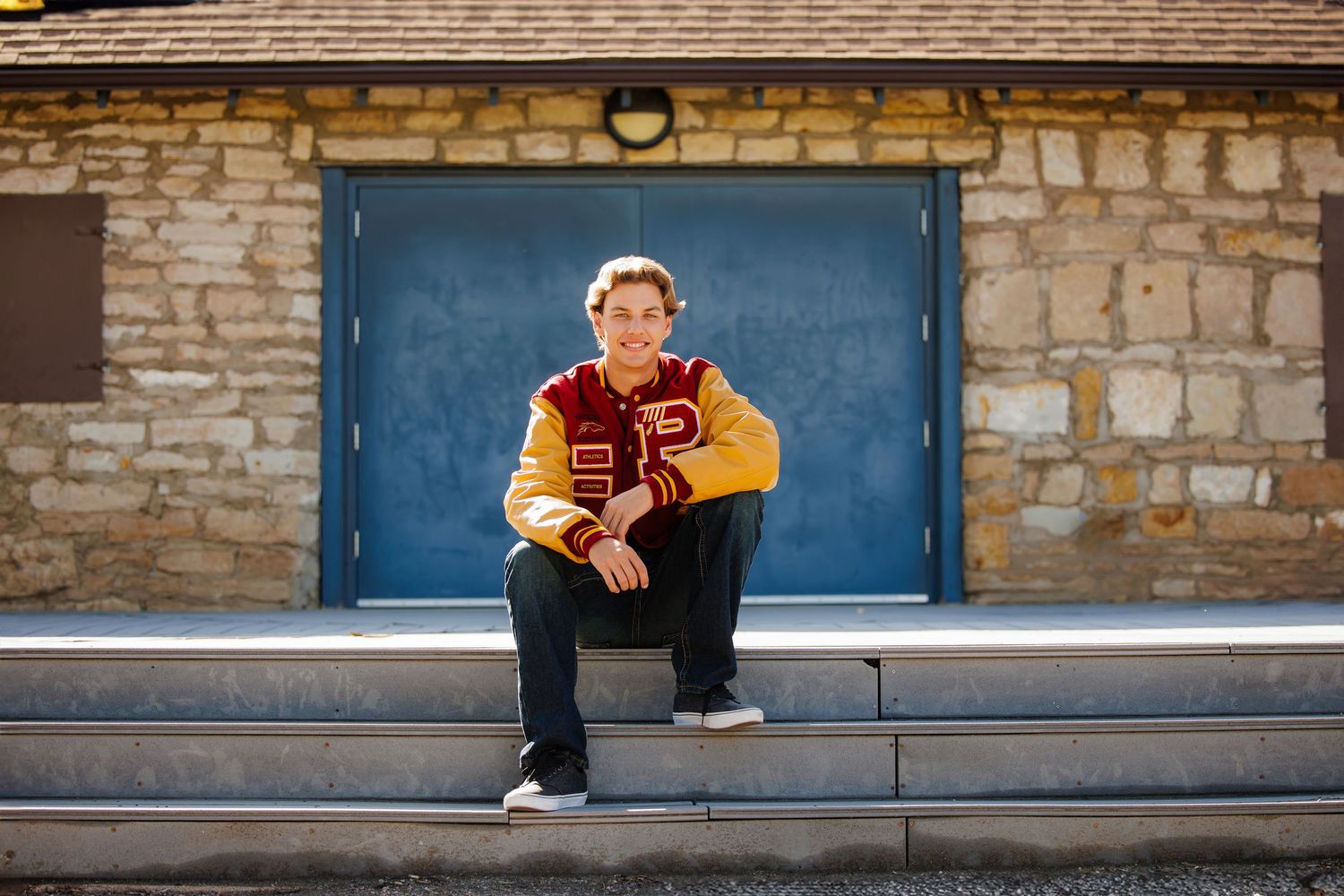 Teen boy in red and gold varsity jacket sitting on stone steps in front of a blue door. Highlands Ranch Colorado Senior Portrait