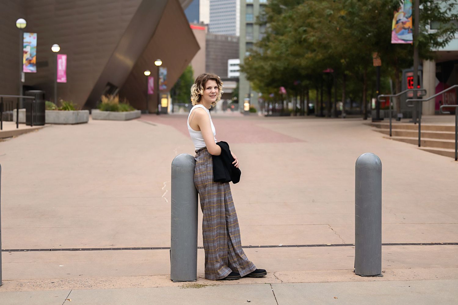 Stylish person in plaid trousers posing between bollards on urban plaza with city buildings behind. Androgynous senior photos Colorado