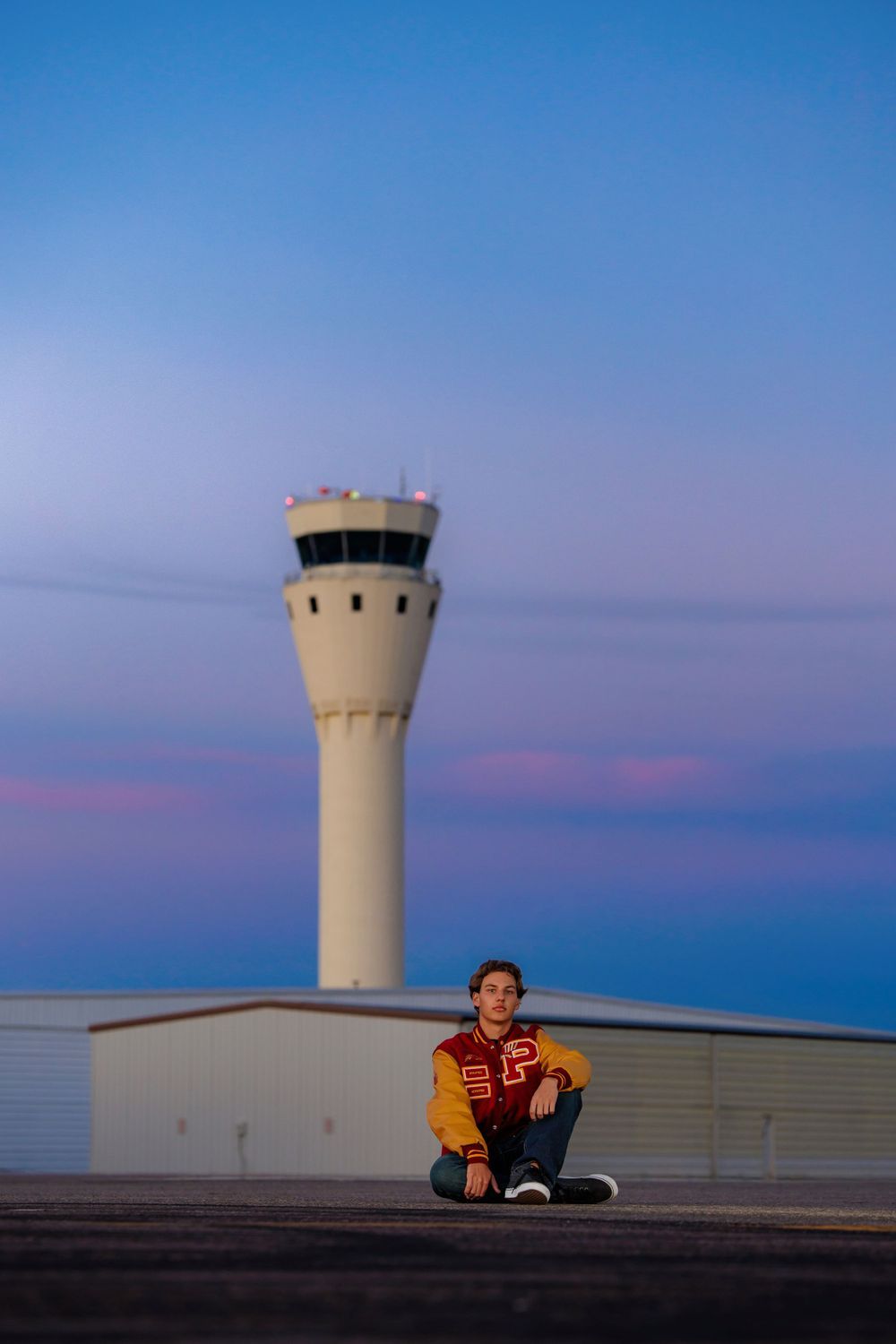 Teen boy in varsity jacket sits on rooftop with an air traffic control tower at dusk. Centennial Colorado Senior Portrait