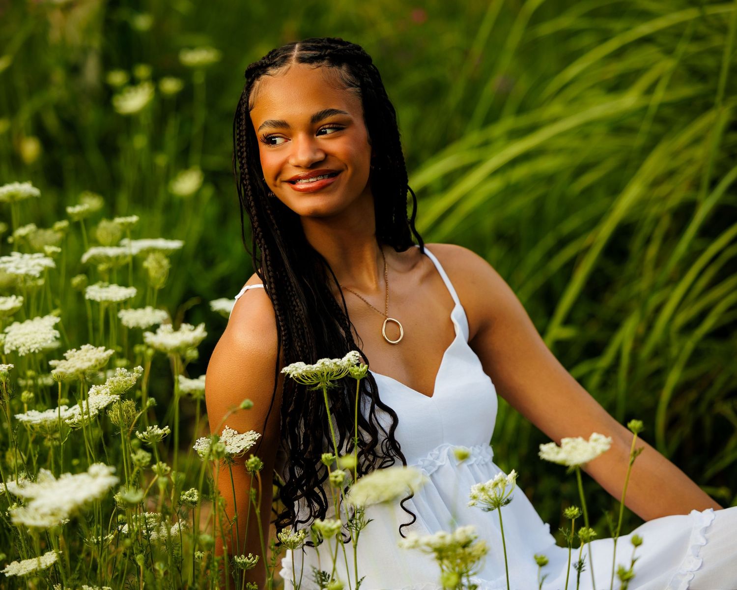 Smiling young woman in white dress sitting among white wildflowers in green field.