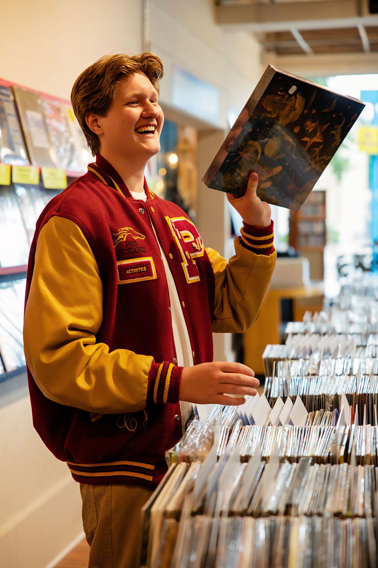 Young man in varsity jacket browsing vinyl records at a music store, smiling while holding an album.