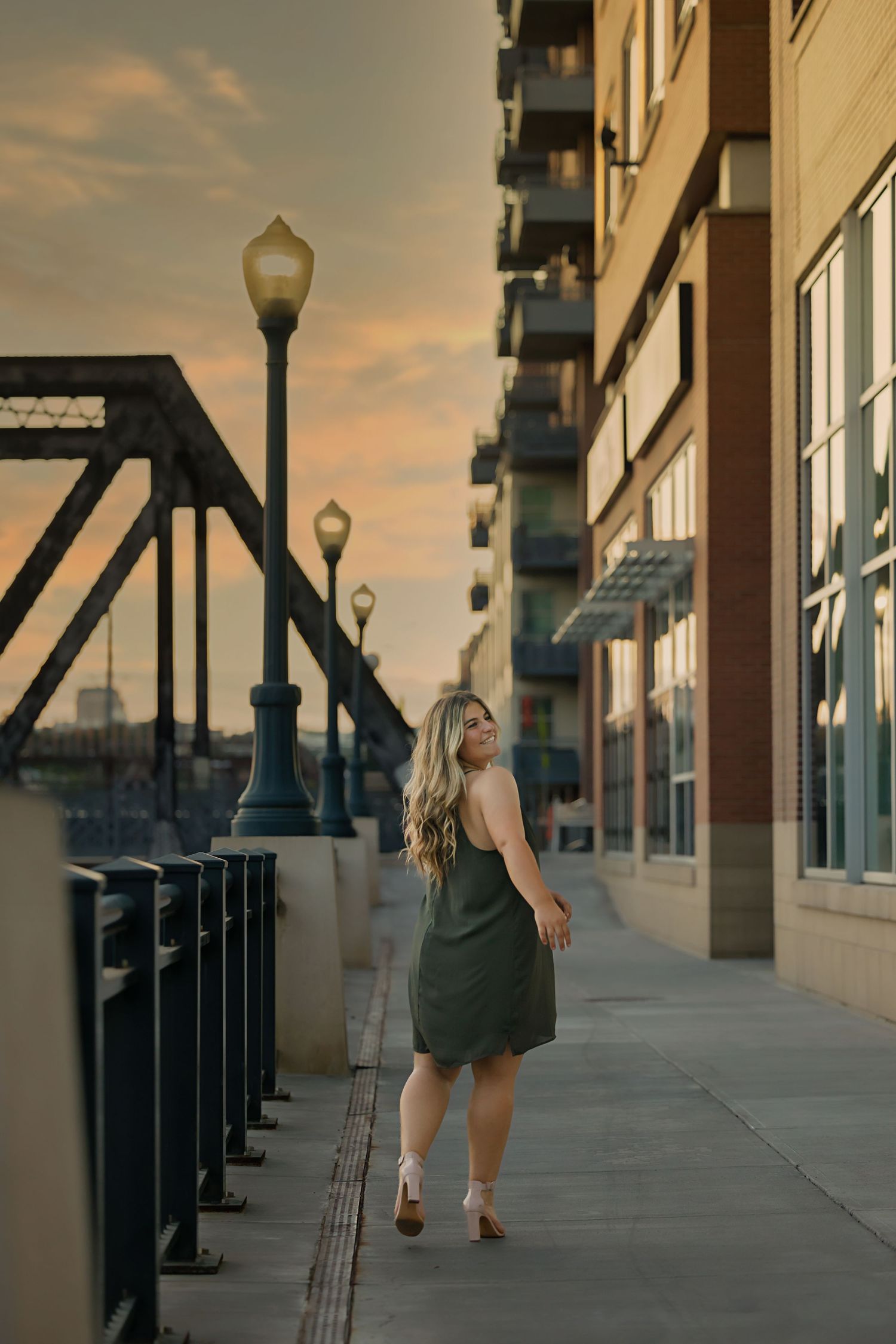 high school senio in green dress walking along urban waterfront promenade at golden hour sunset with glowing street lamps in Downtown Denver