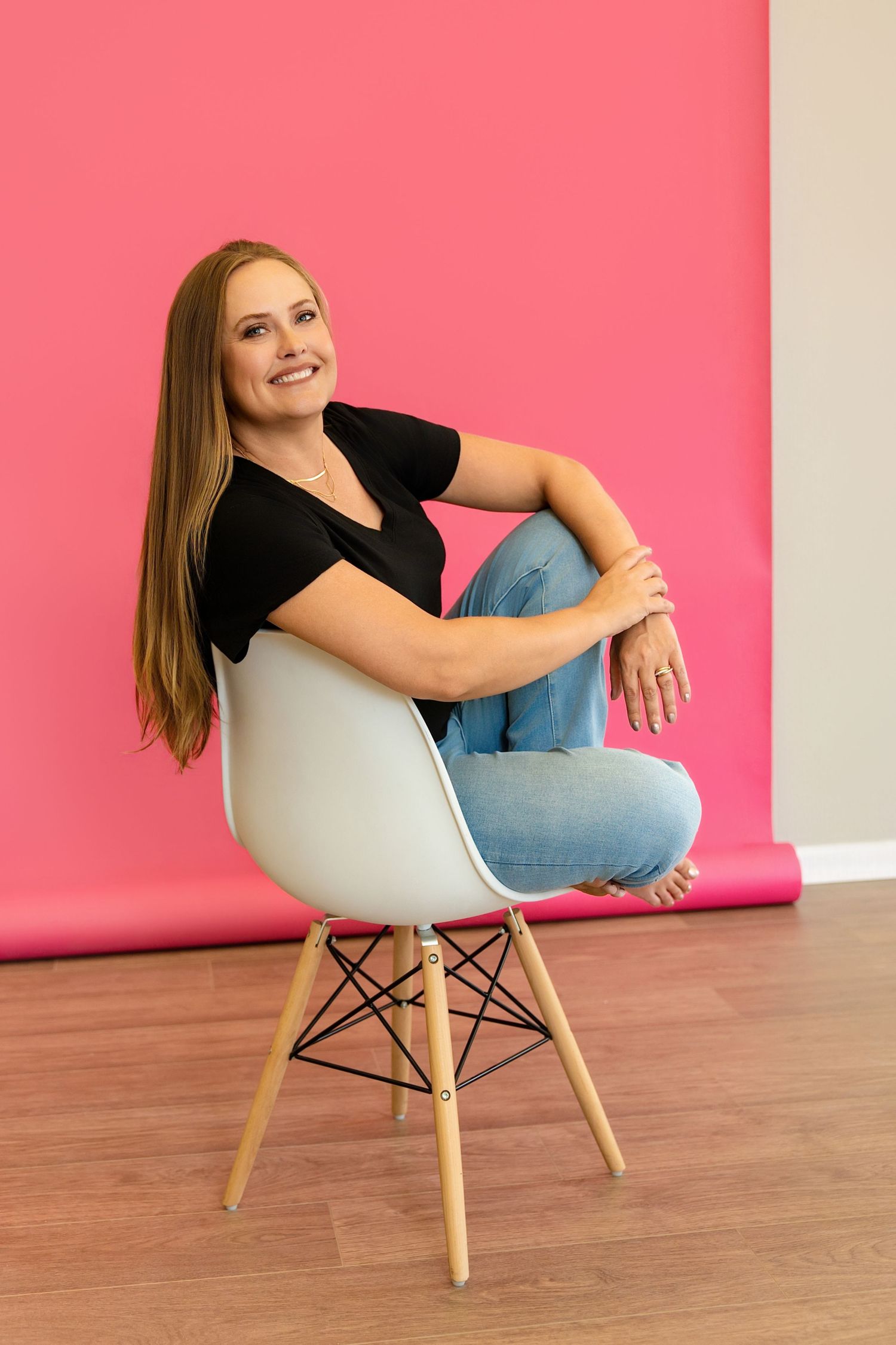 Melissa Nansen: long brown hair sitting casually in a white chair against a vibrant pink background in her Douglas County Portrait Studio