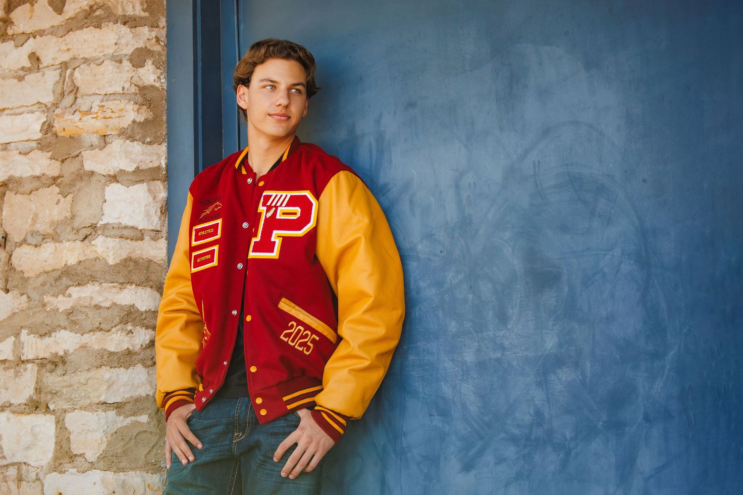 Young man in red and gold letterman jacket posing against blue wall for senior portrait. Aurora Colorado Senior Portrait