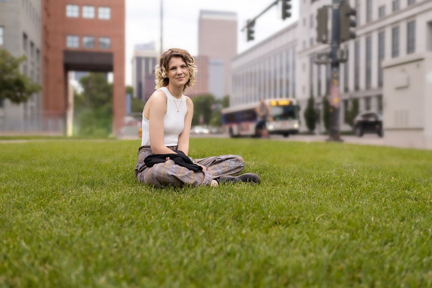 Human in white tank top sitting cross-legged on green grass lawn in urban city setting.