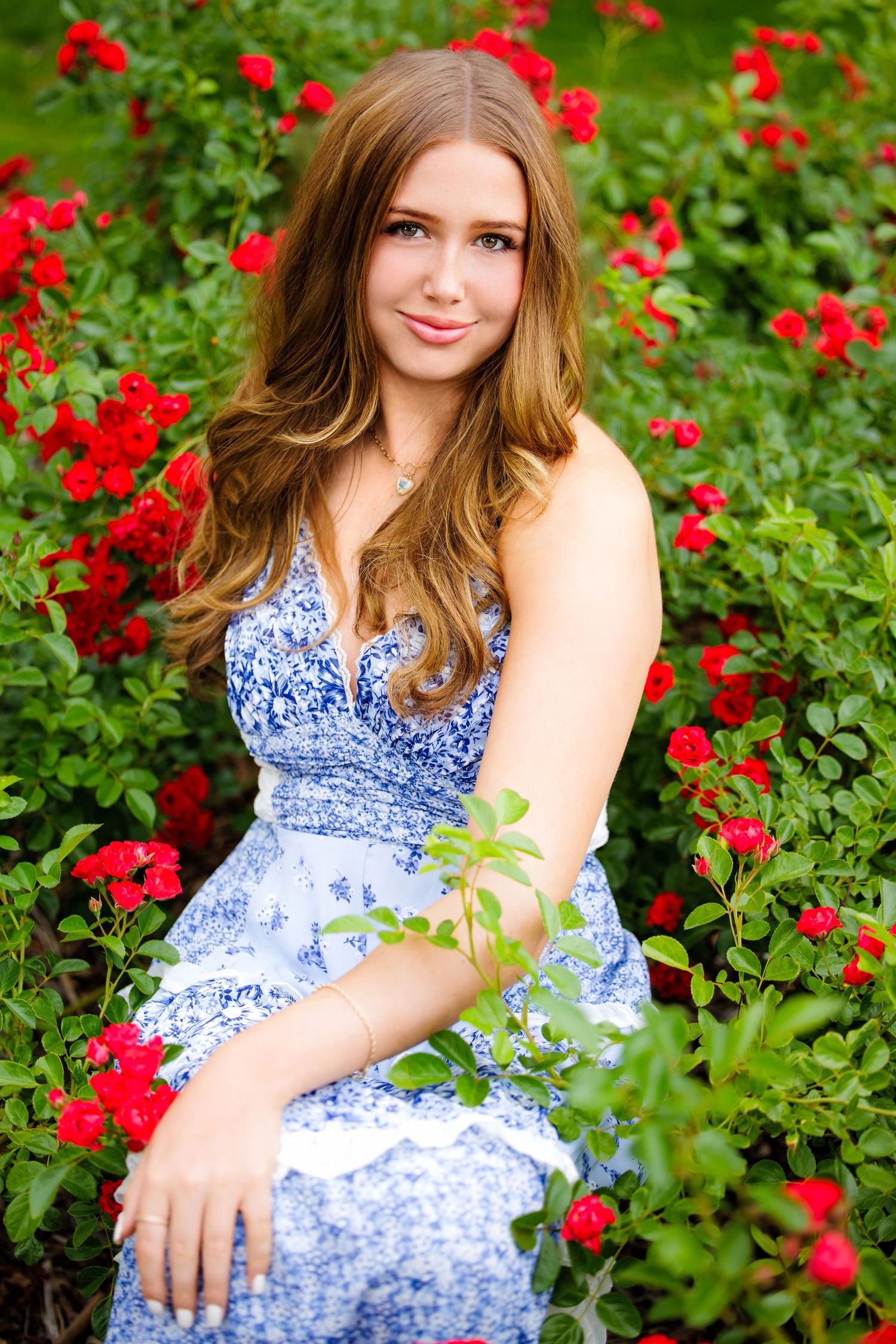 Young woman in blue floral dress sitting among vibrant red roses in a lush green garden.