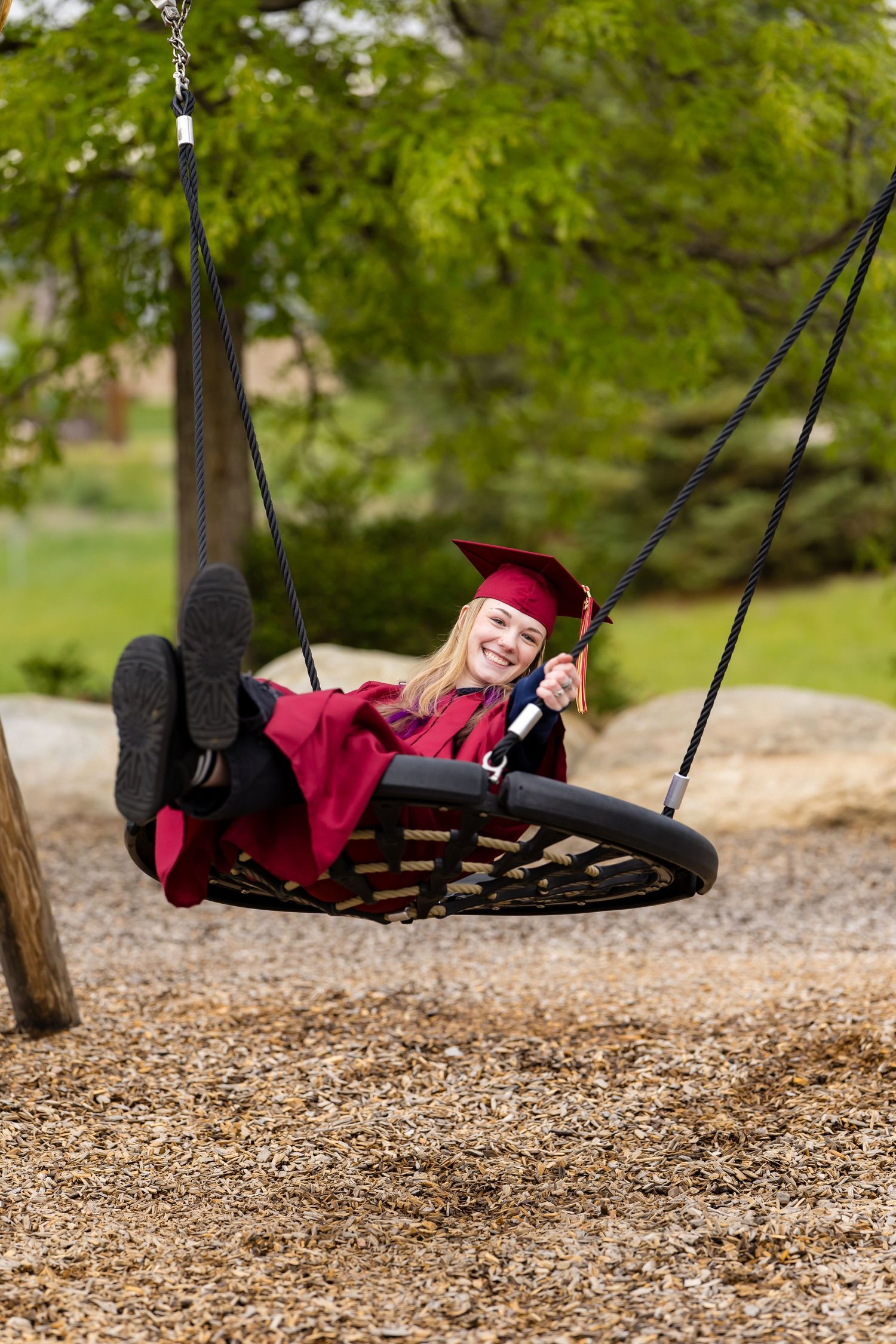 Happy female graduate in red cap and gown swinging on playground swing with diploma.