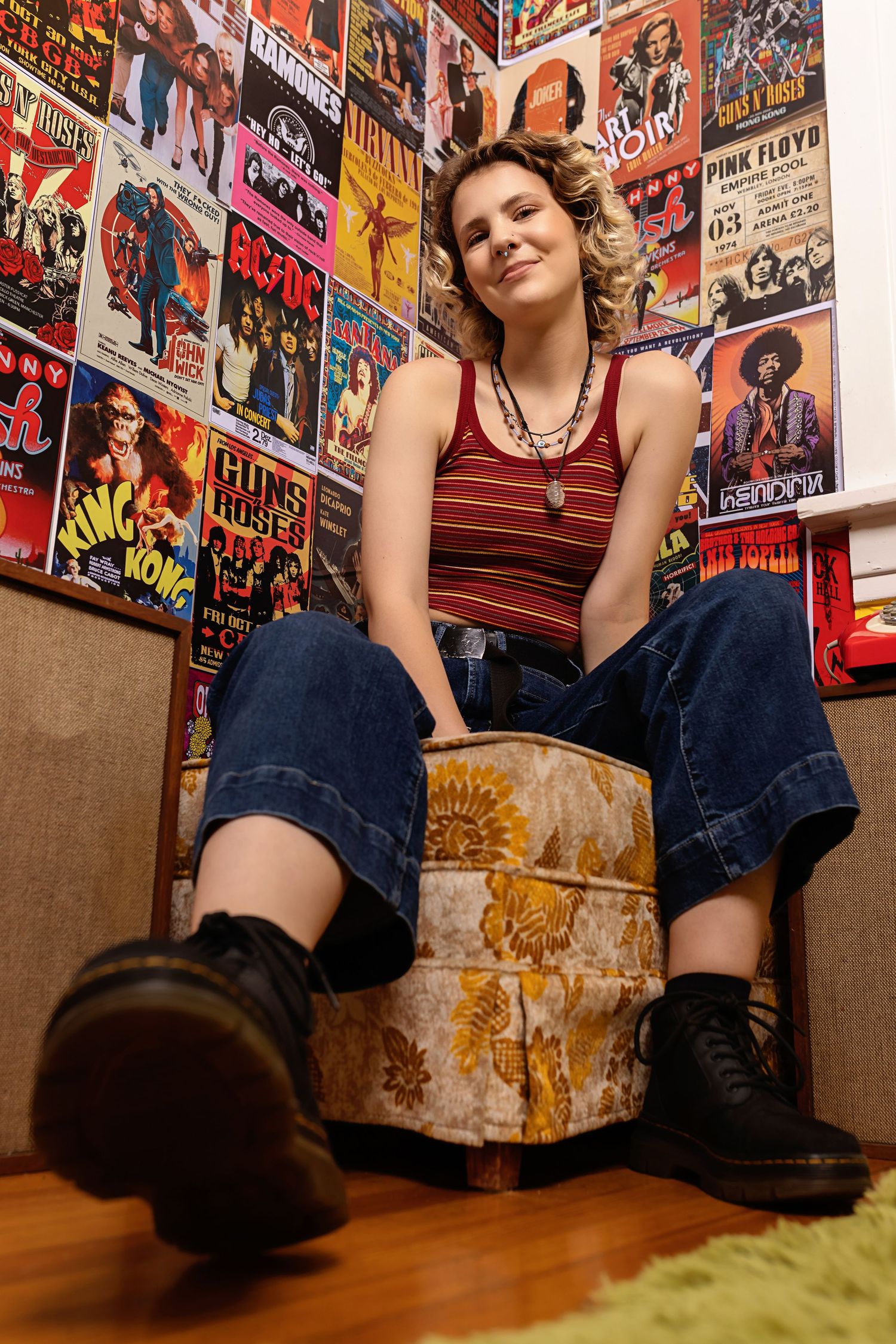Teen in striped tank top and jeans sits on vintage ottoman surrounded by rock music posters posing for senior photos Colorado