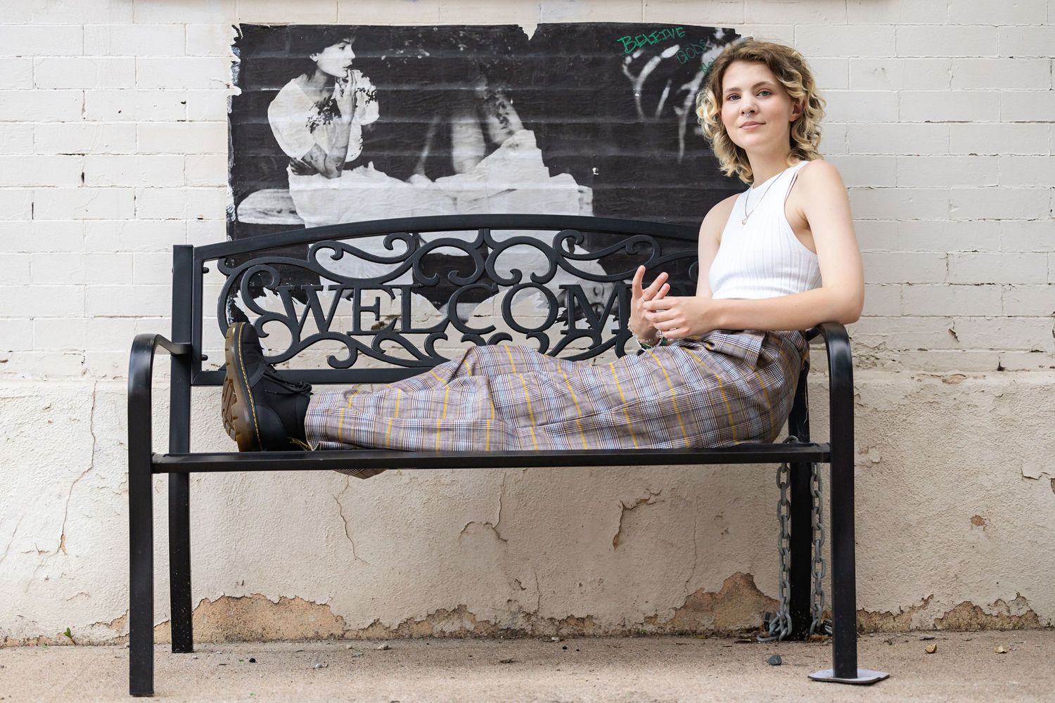 Androgynous Teen in plaid wide-leg pants relaxing on decorative welcome bench against white brick wall. Denver Senior Photos