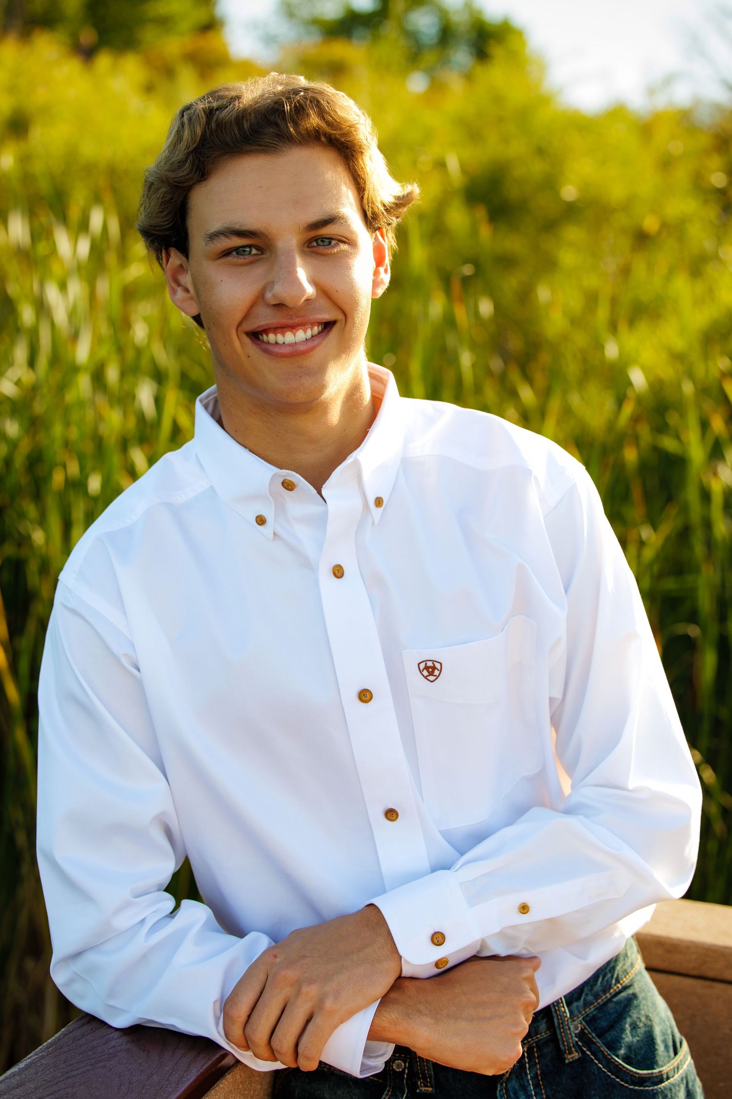Smiling teenage boy in white Ariat shirt seated outdoors with green grass background. Littleton Colorado Senior Portrait