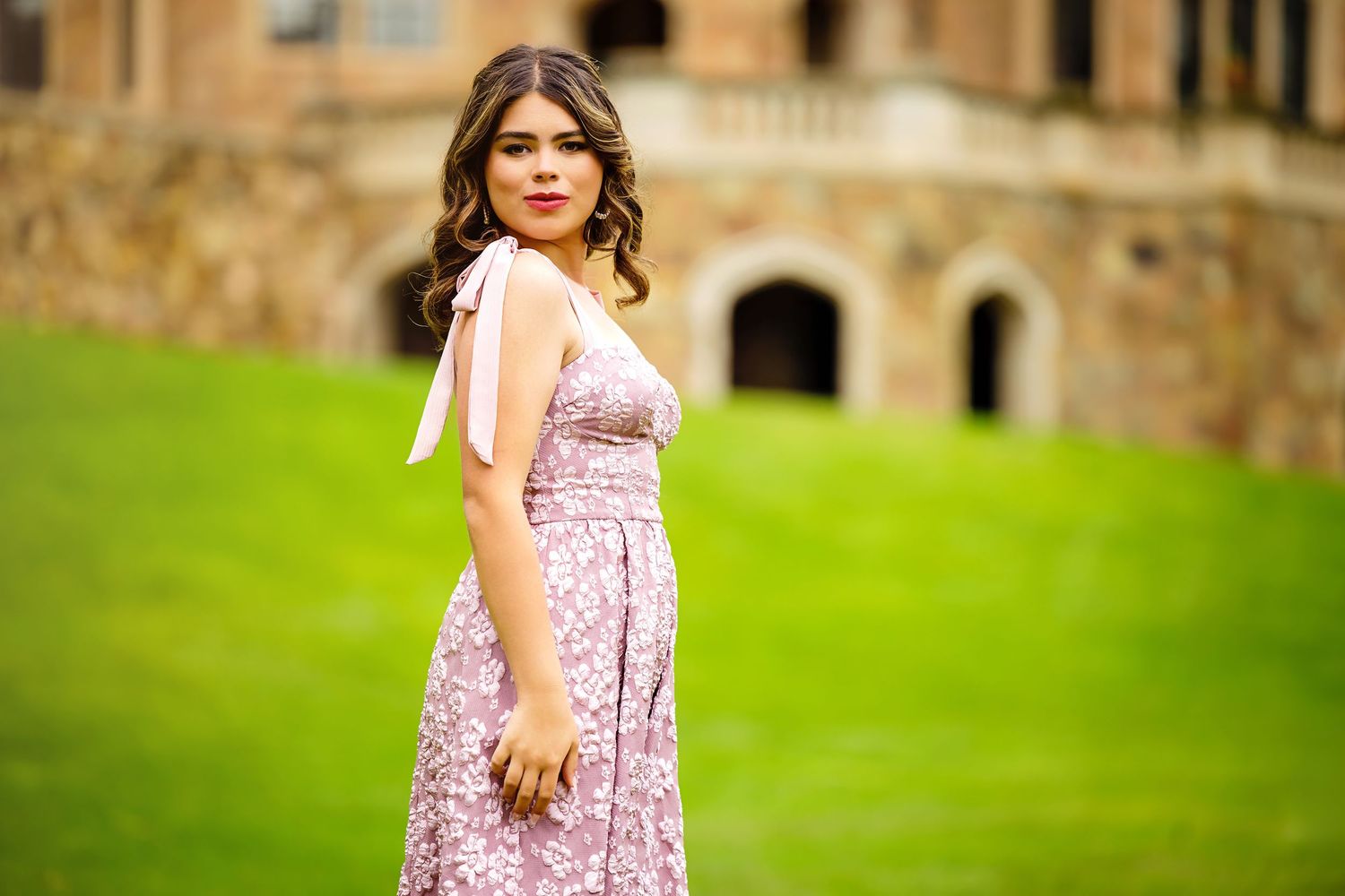 Young woman in pink floral midi dress with bow shoulder tie poses on green lawn before historic stone building for Denver Senior pictures
