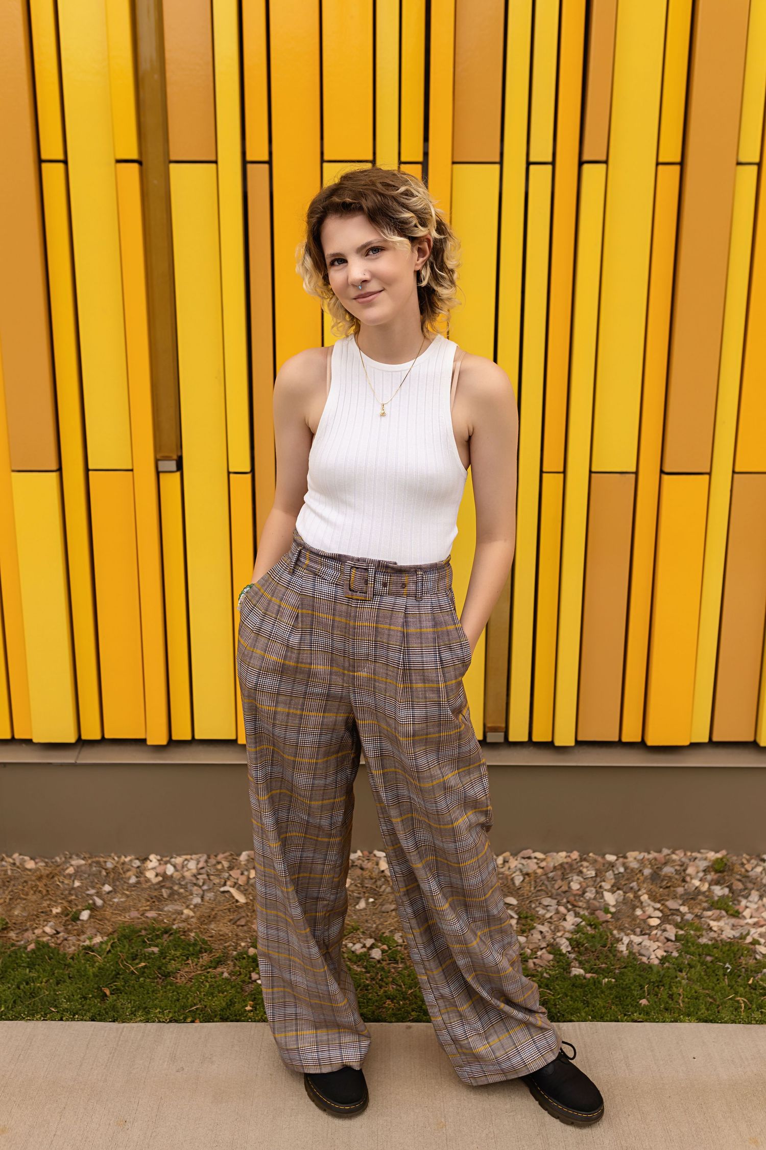 Person in white tank top and plaid wide-leg pants stands confidently before yellow striped wall.