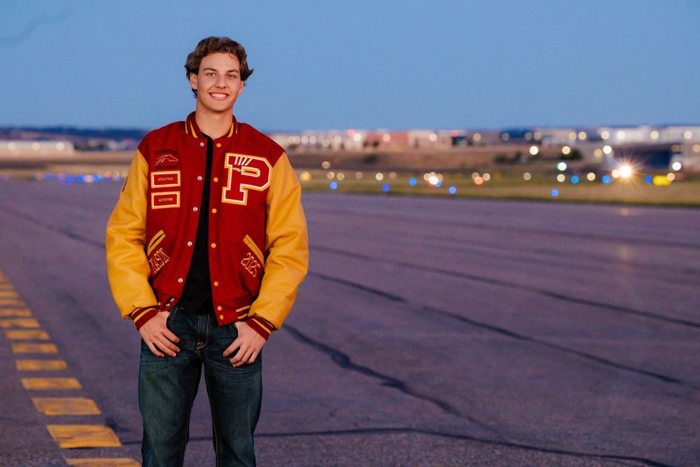 Young man in red gold letterman jacket standing on airport runway at twilight with bokeh lights behind. Lonetree Colorado Senior Portrait