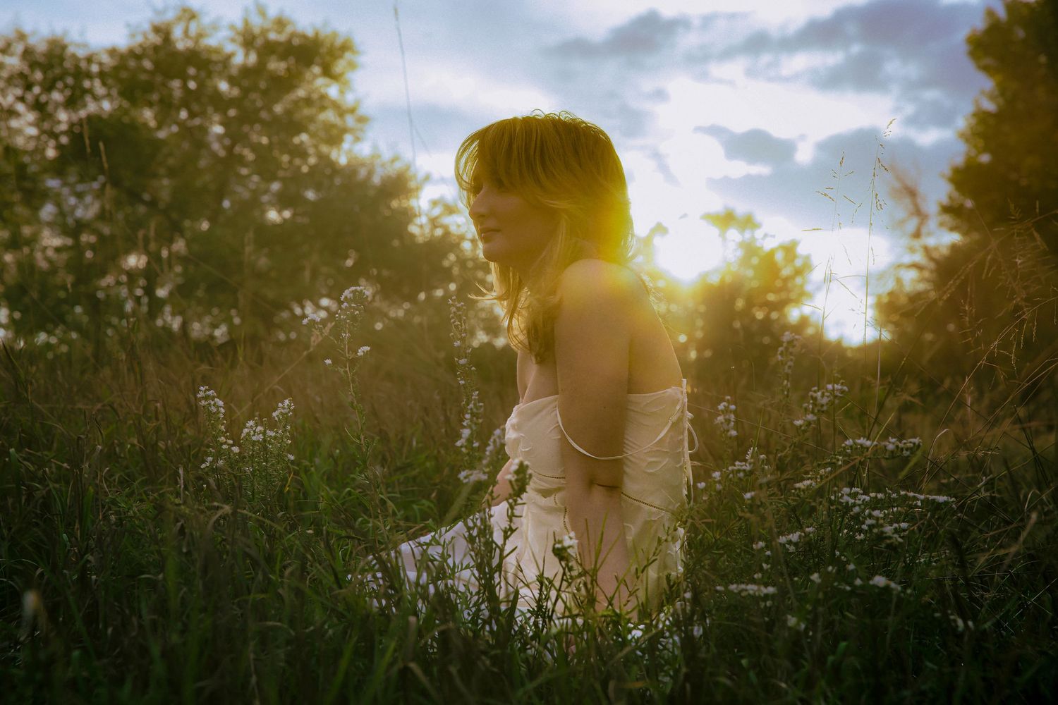 Woman in white dress sitting in tall grass during golden hour sunset with glowing backlight and cloudy sky.