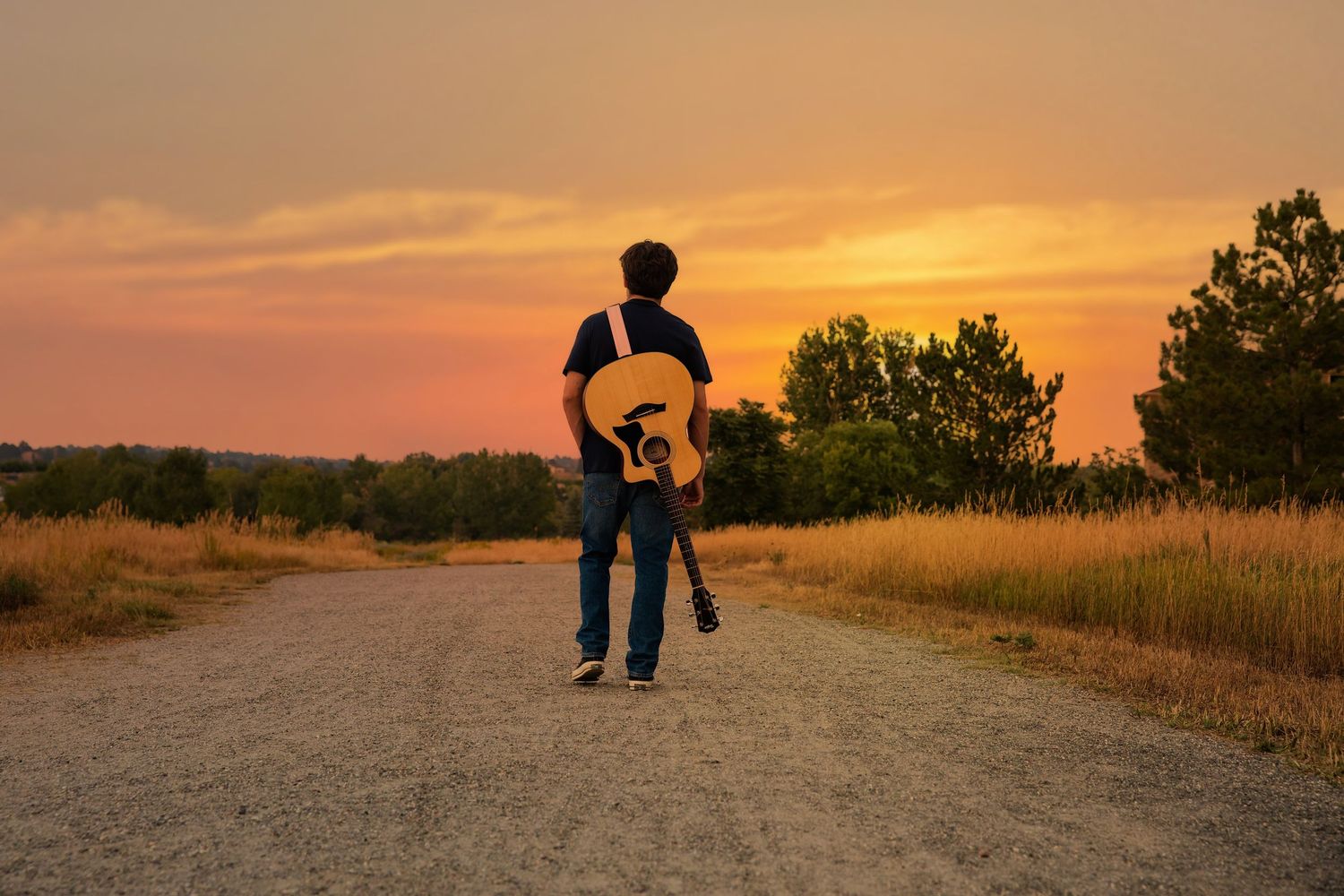 Person carrying guitar walking on gravel road toward stunning orange sunset.