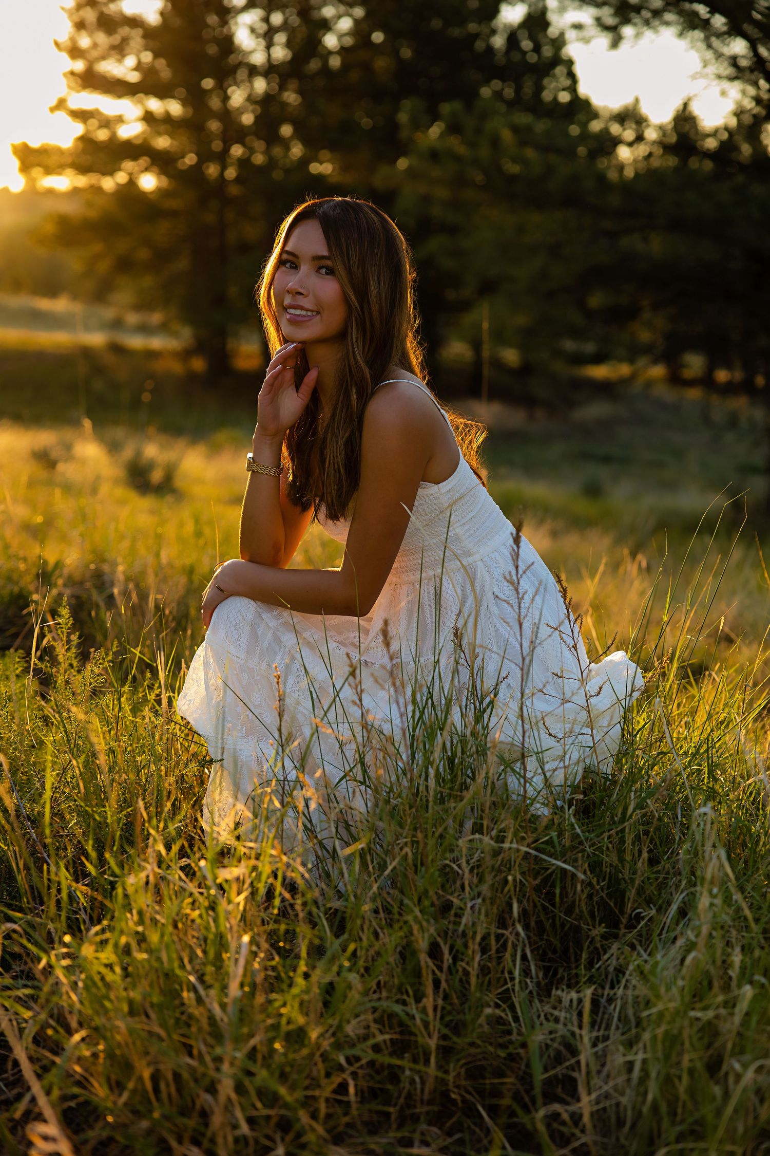 Woman in a white sundress crouching in tall golden grass at sunset, smiling softly with warm sunlight filtering through pine trees.