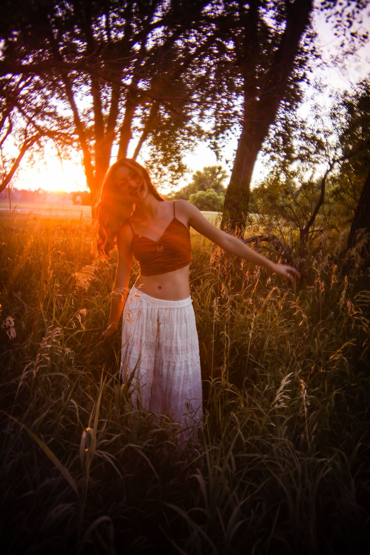 Woman in dark crop top and white skirt with arms outstretched in tall grass field during vibrant golden sunset for Colorado Senior pictures