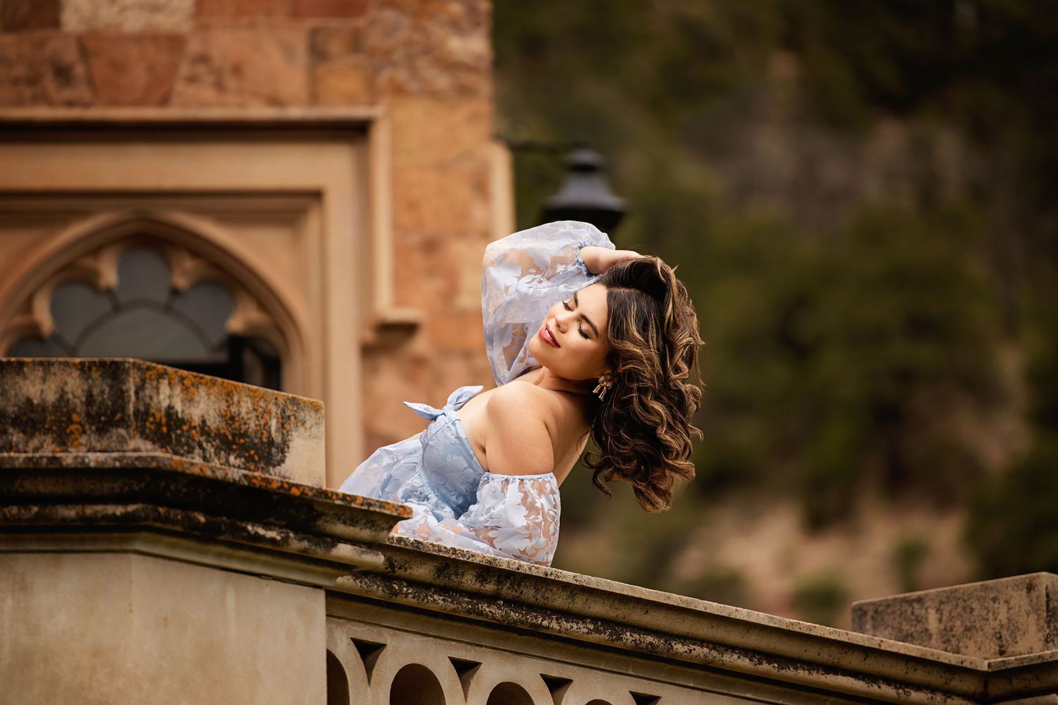 Elegant woman in floral blue dress leaning back gracefully on stone balcony at historic building Paker CO High School Senior pictures