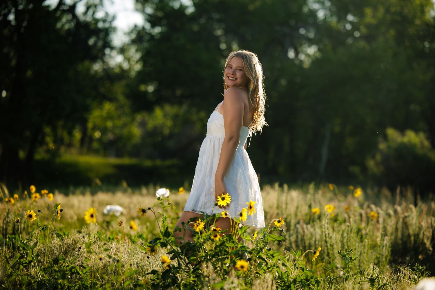 Young blonde woman in white dress standing in a sunny wildflower meadow, smiling over her shoulder.