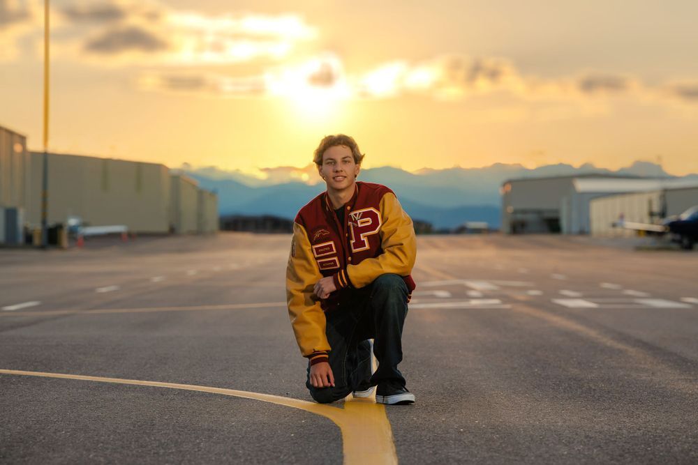 Teen in varsity jacket crouches on yellow tarmac line during golden sunset outdoors. Arapahoe County Colorado Senior Portrait