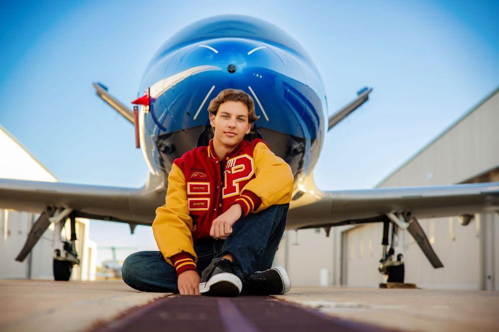 Teen in letterman jacket sits on runway in front of blue aircraft nose at airport. Ponderosa Colorado Senior Portrait