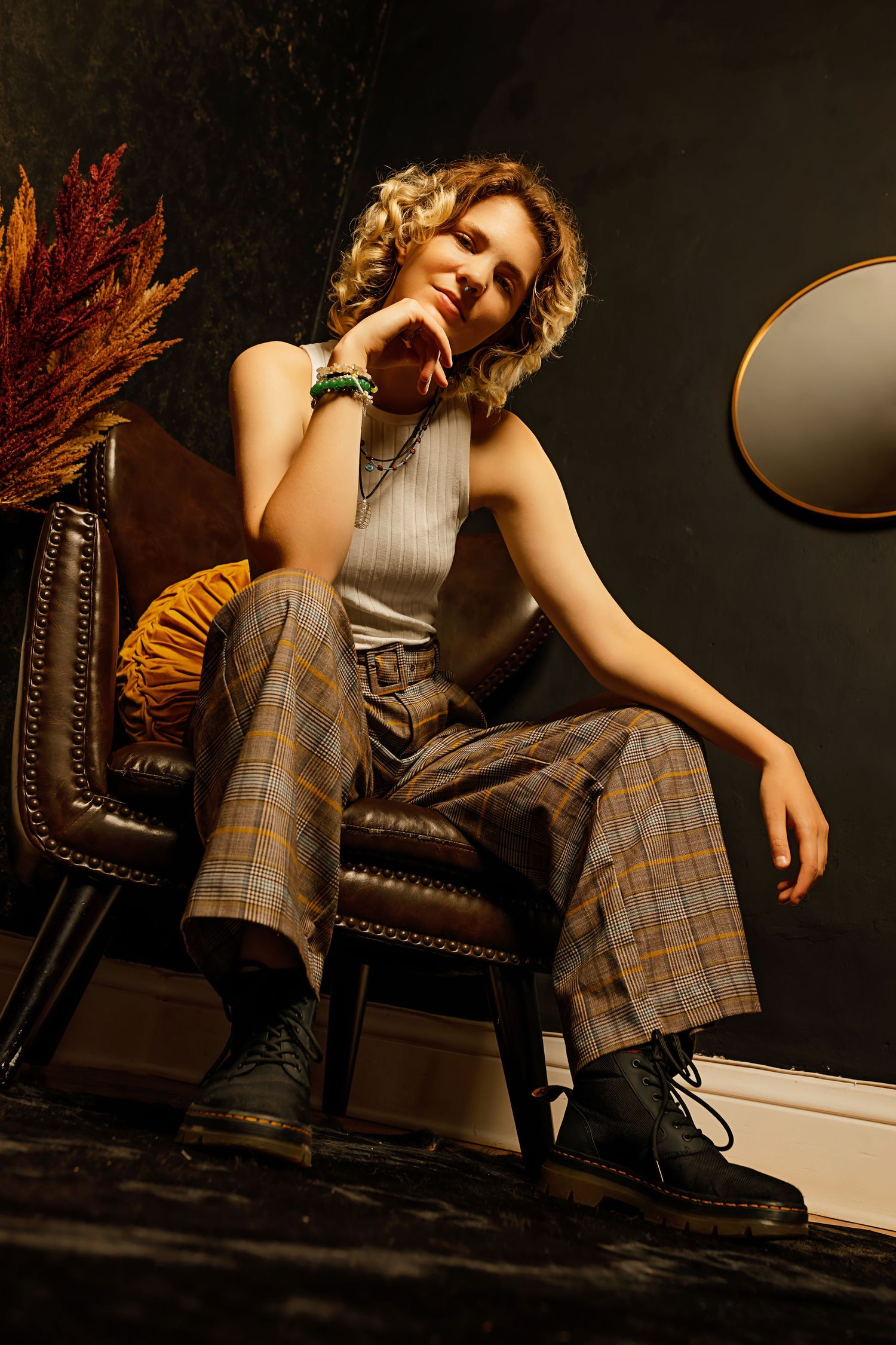 Person with curly blonde hair in plaid wide-leg pants sits in leather chair in dramatic dark setting.