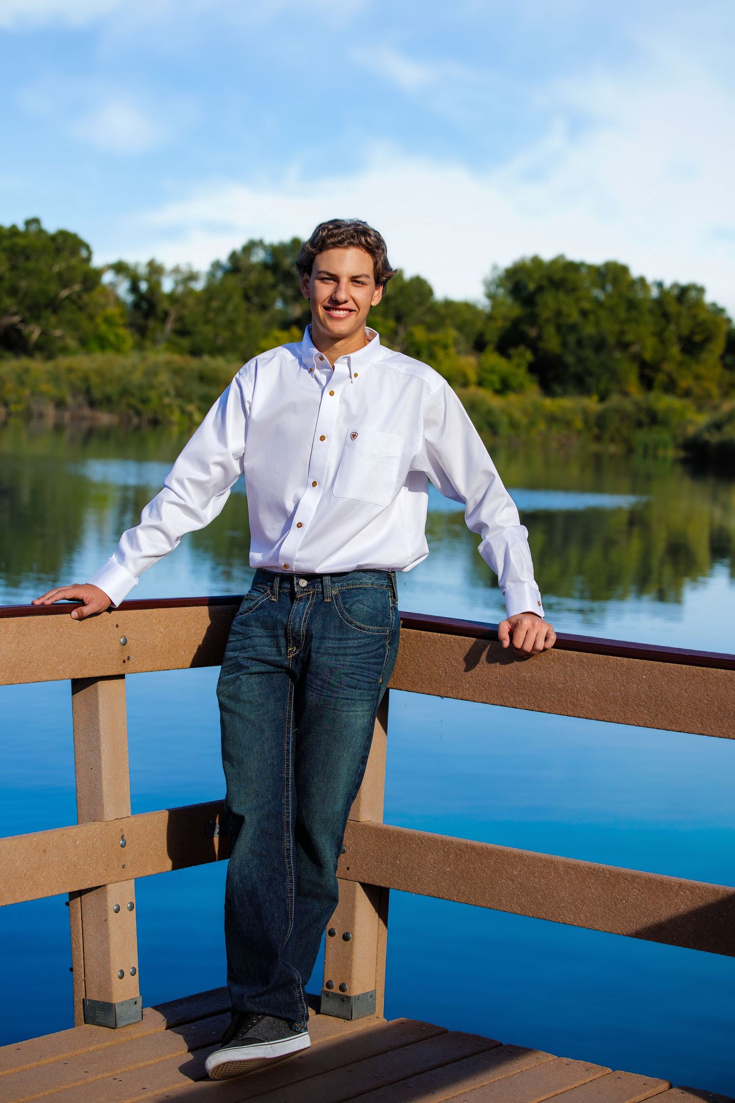 Smiling teen boy in white shirt leaning on wooden bridge railing with lake backdrop.  Denver Colorado Senior Portrait