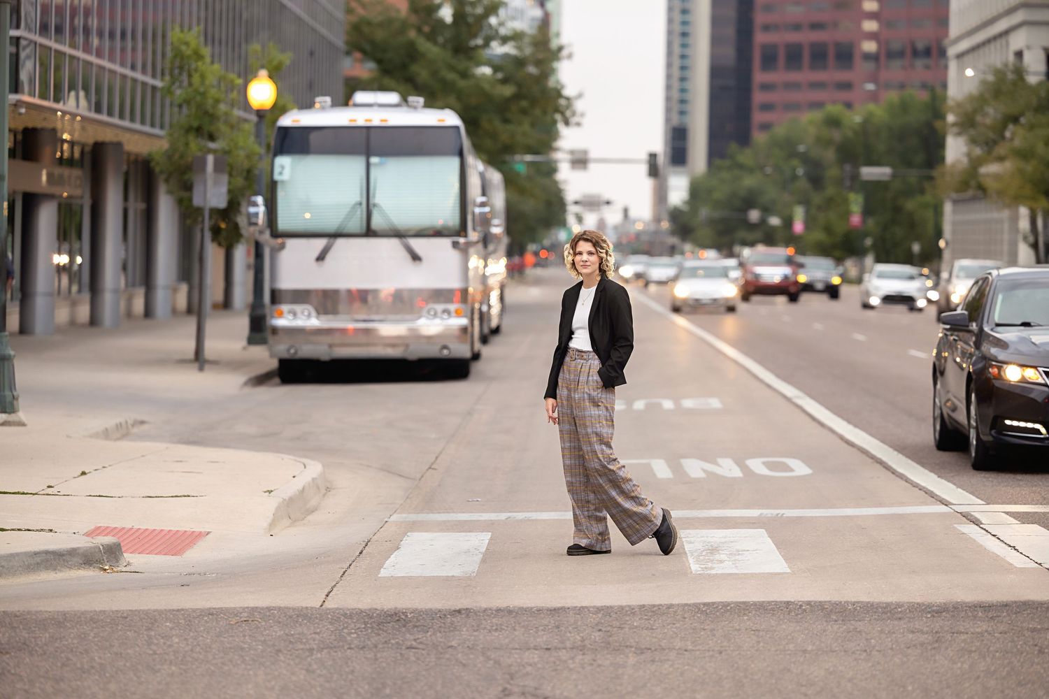 Teen in wide-leg plaid pants crossing urban street with city bus in background. Androgynous Senior photos Douglas County Colorado