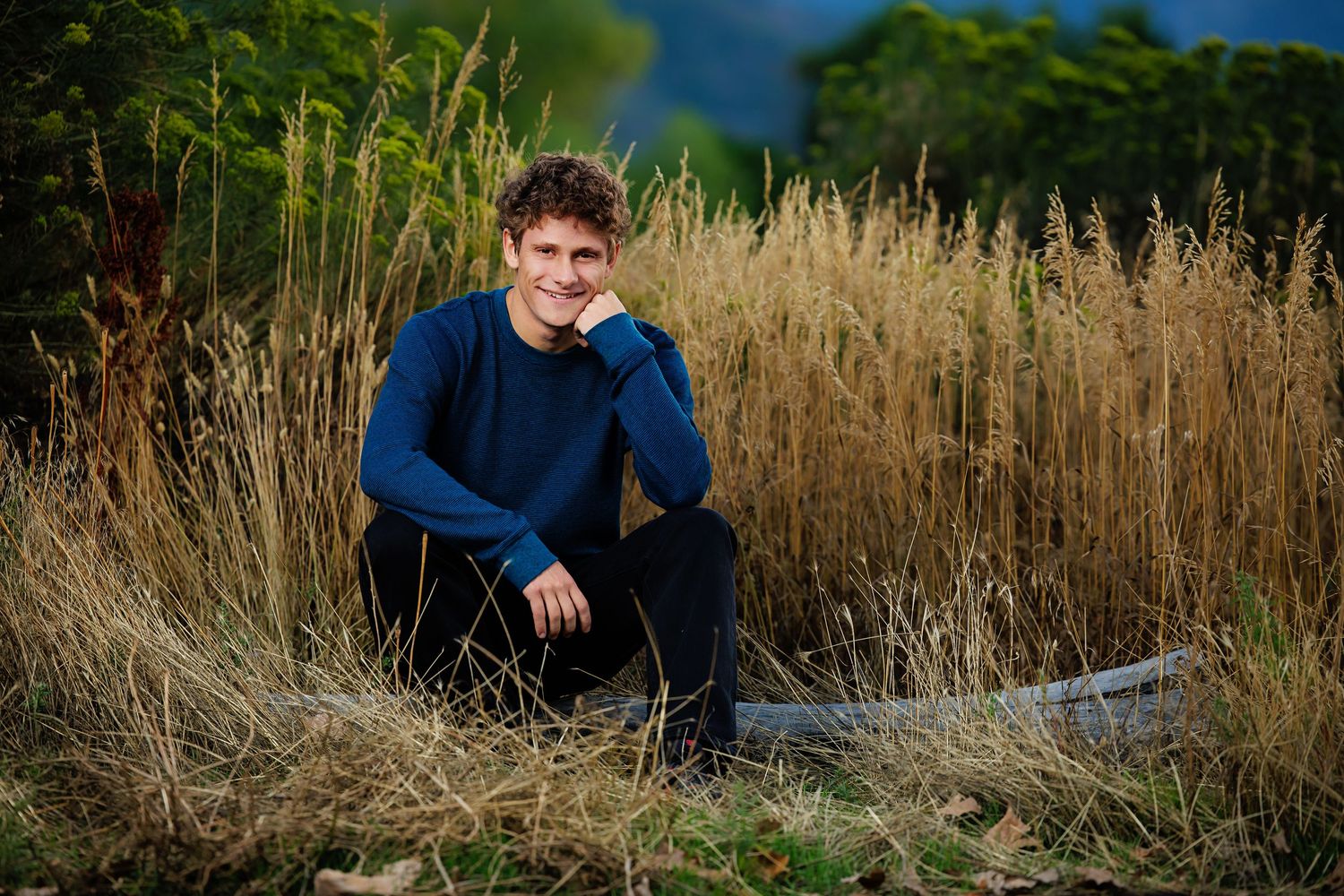 Young man in blue sweater sitting outdoors among tall golden grass, smiling naturally in a scenic nature setting.