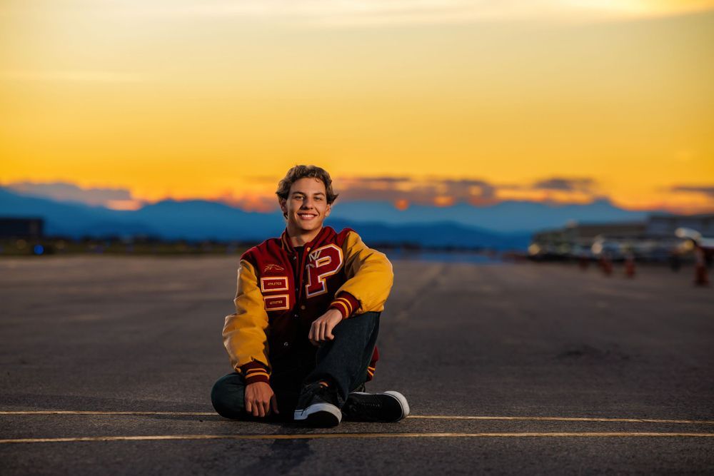 Smiling teen in red and gold varsity jacket sits on tarmac at golden hour sunset. Ponderosa Colorado Senior Portrait