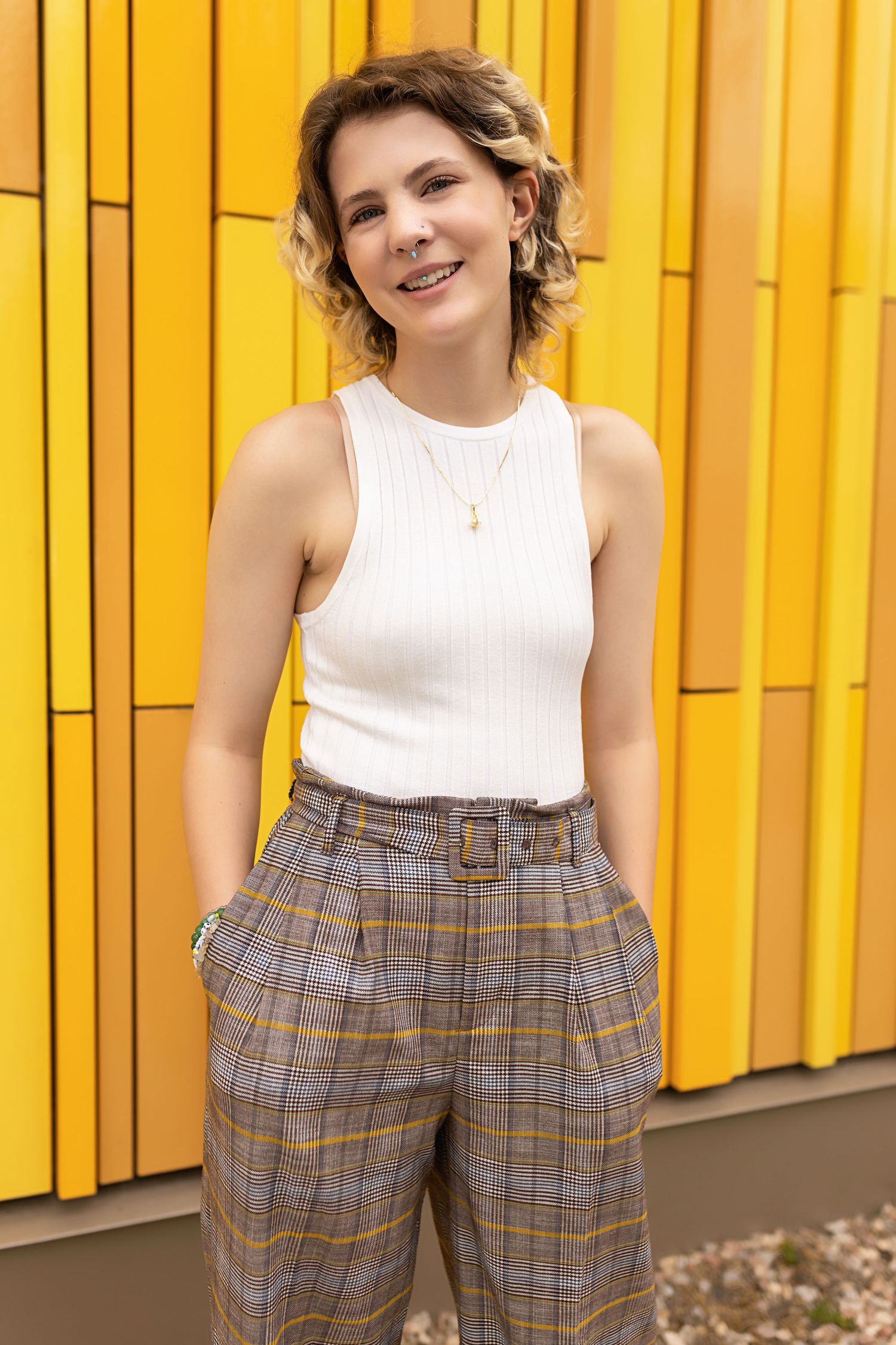 Androgynous teen in white sleeveless top and belted plaid pants stands before bright yellow wall Downtown Denver senior photos