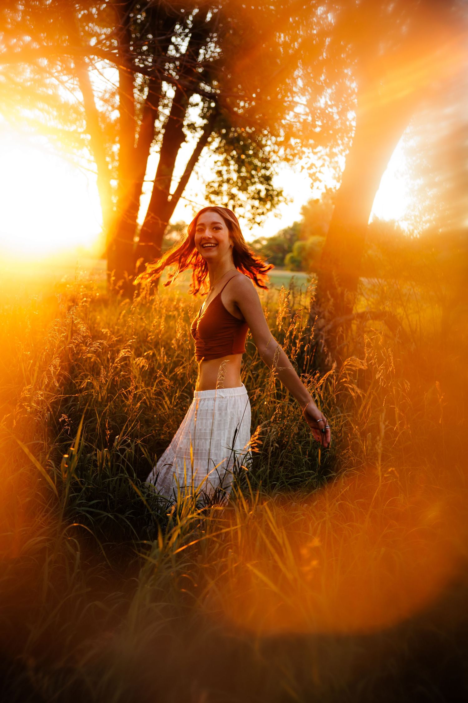 Joyful woman in brown crop top and white skirt dancing in golden sunset light among tall grass and trees for Elizabeth CO Senior photos