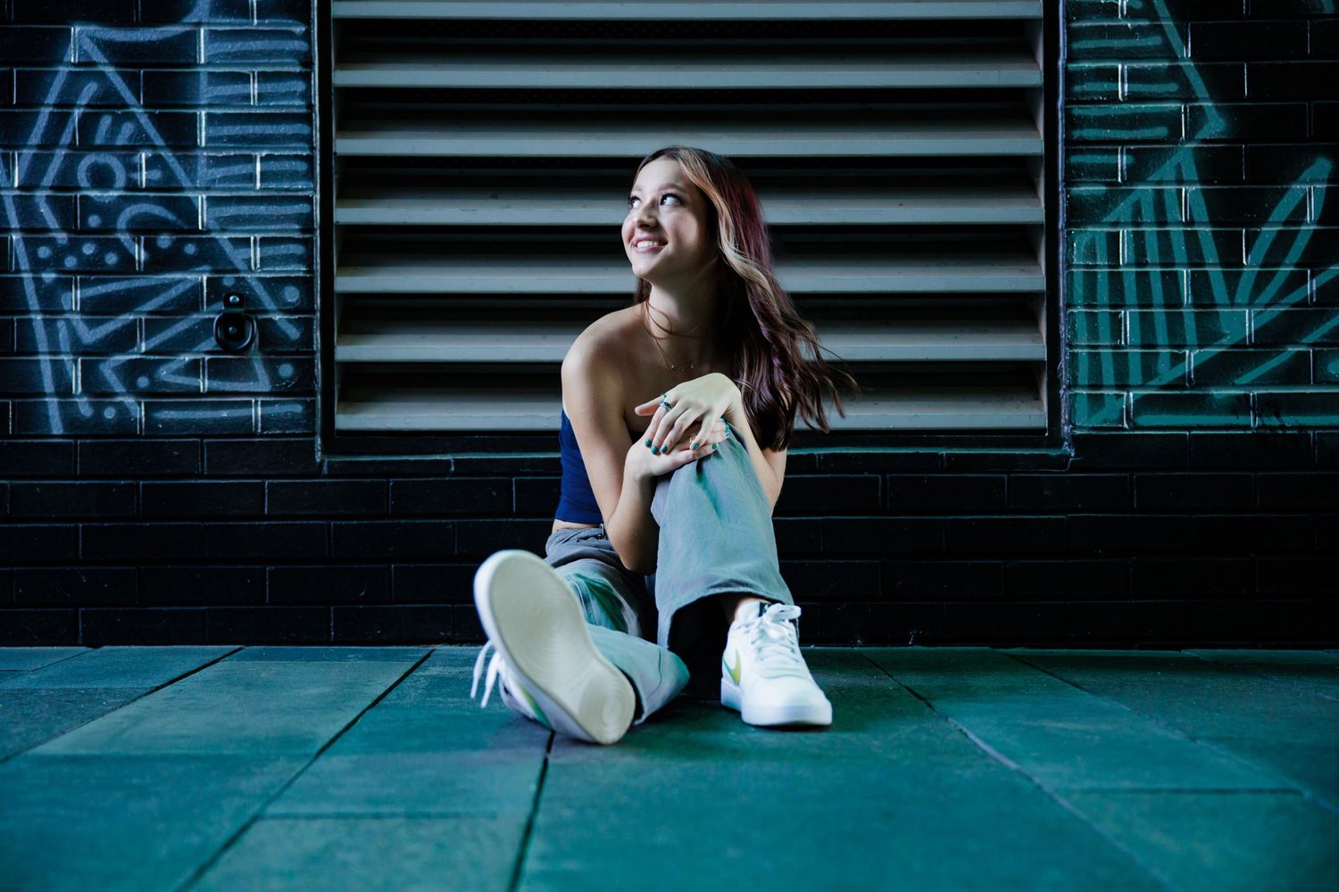 Young woman sitting on ground laughing outdoors at night near graffiti wall for Downtown Denver CO Senior photos