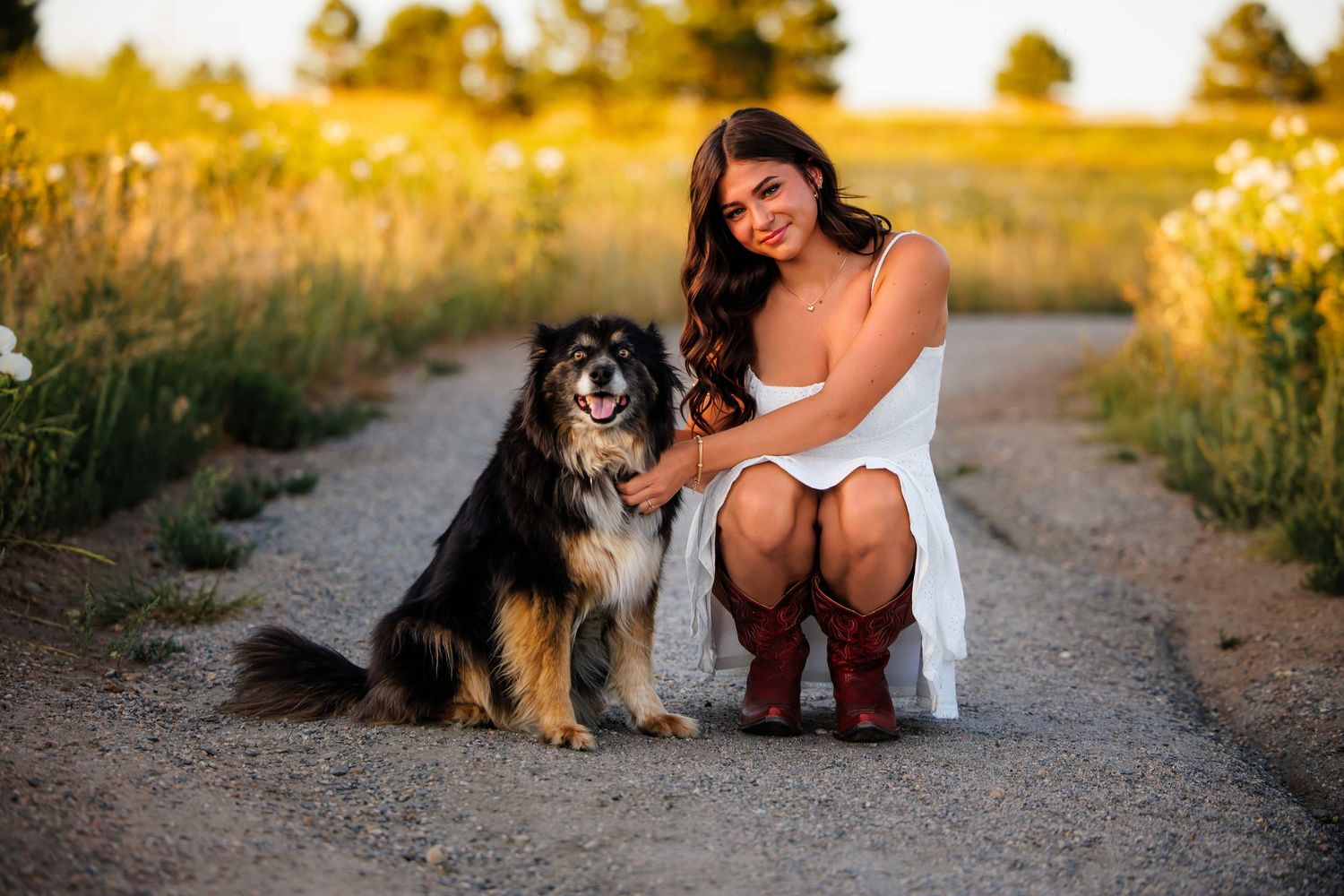 Young woman in white dress and cowboy boots crouches beside fluffy dog on rural dirt path at golden hour.